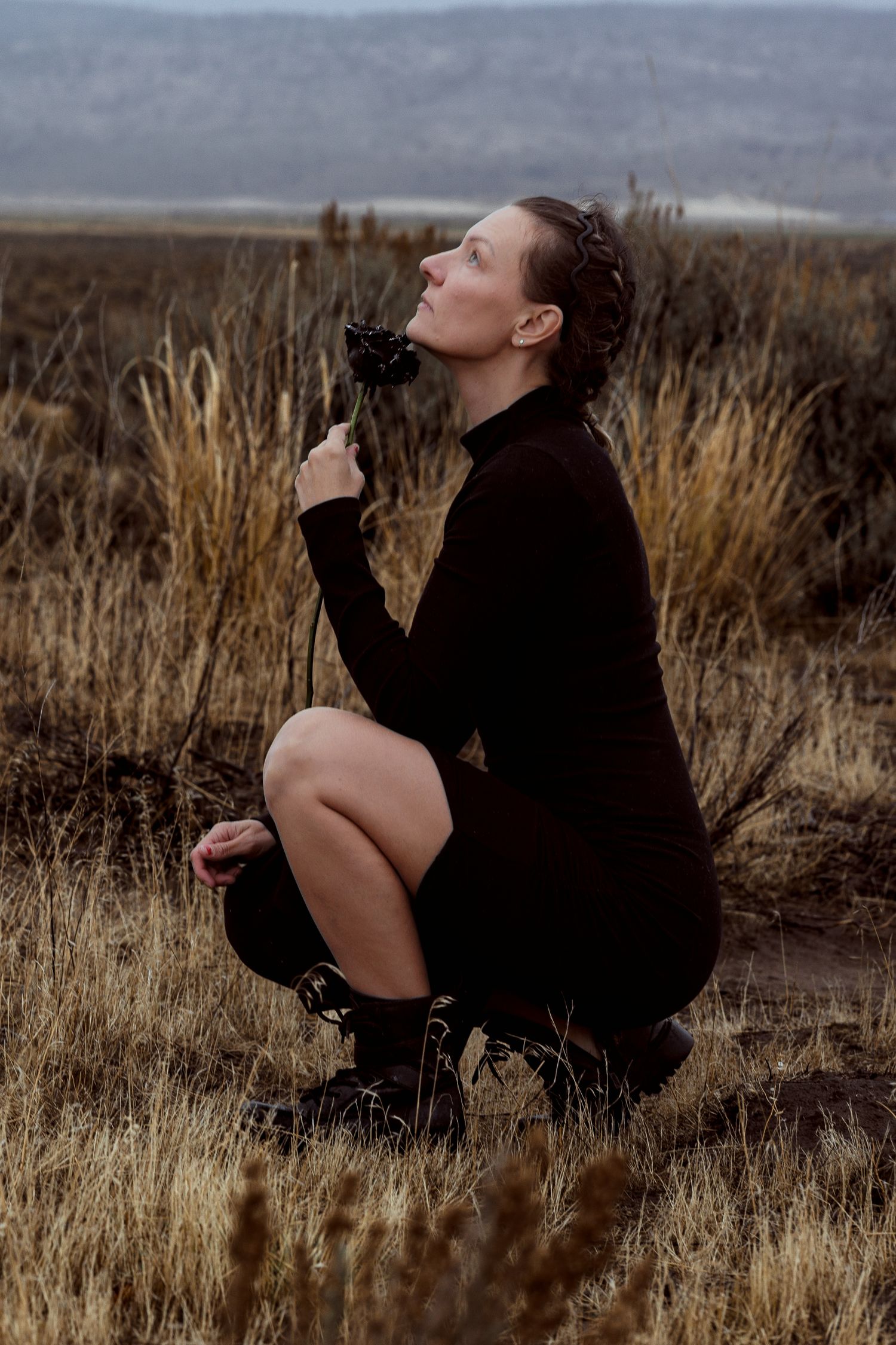 Woman in black turtleneck dress crouching in dry grassland field, holding a dark flower while gazing upward.