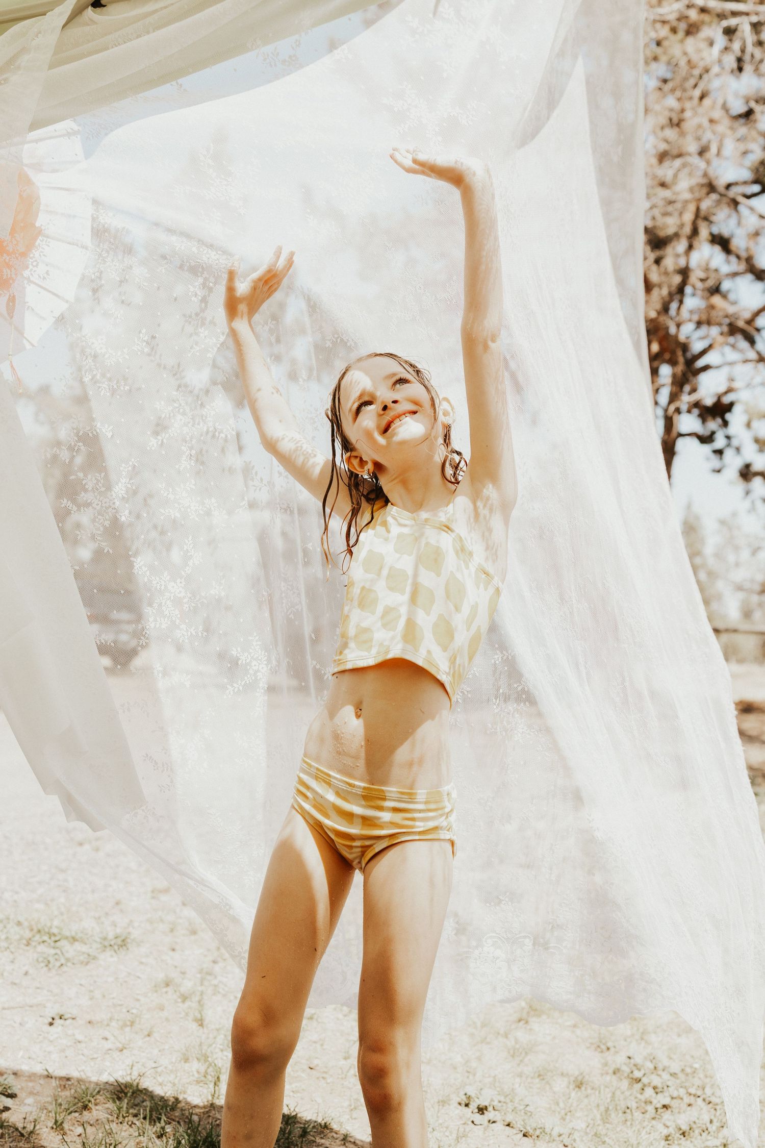 A joyful girl in a yellow bikini smiles with arms raised as white fabric billows around her.