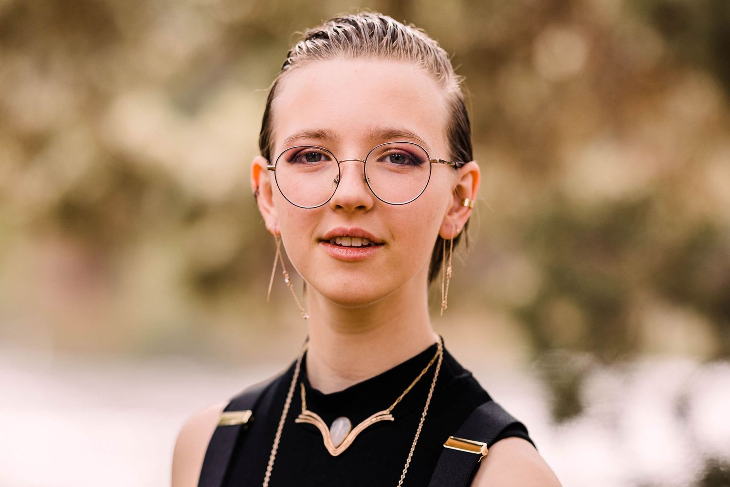 Young person with slicked back hair wearing round glasses, black outfit, and layered gold jewelry outdoors.