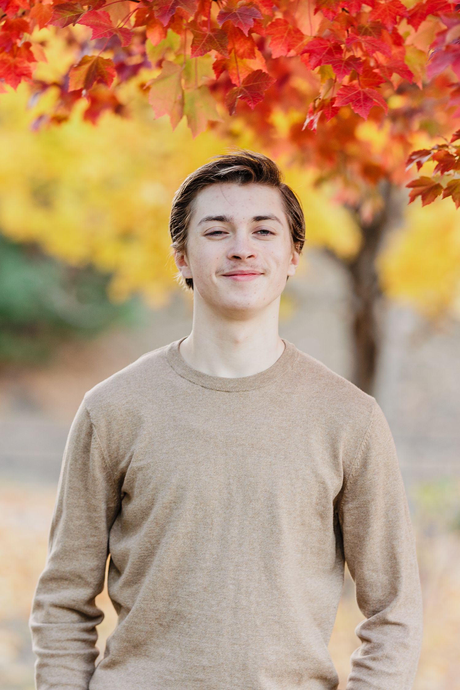 Smiling teenage boy in a beige sweater standing outdoors with vibrant red and orange autumn foliage.