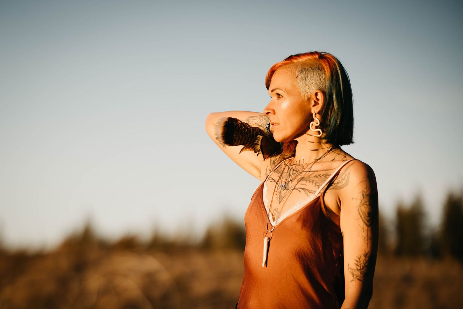 Tattooed woman in rust slip dress gazes into distance outdoors at golden hour.
