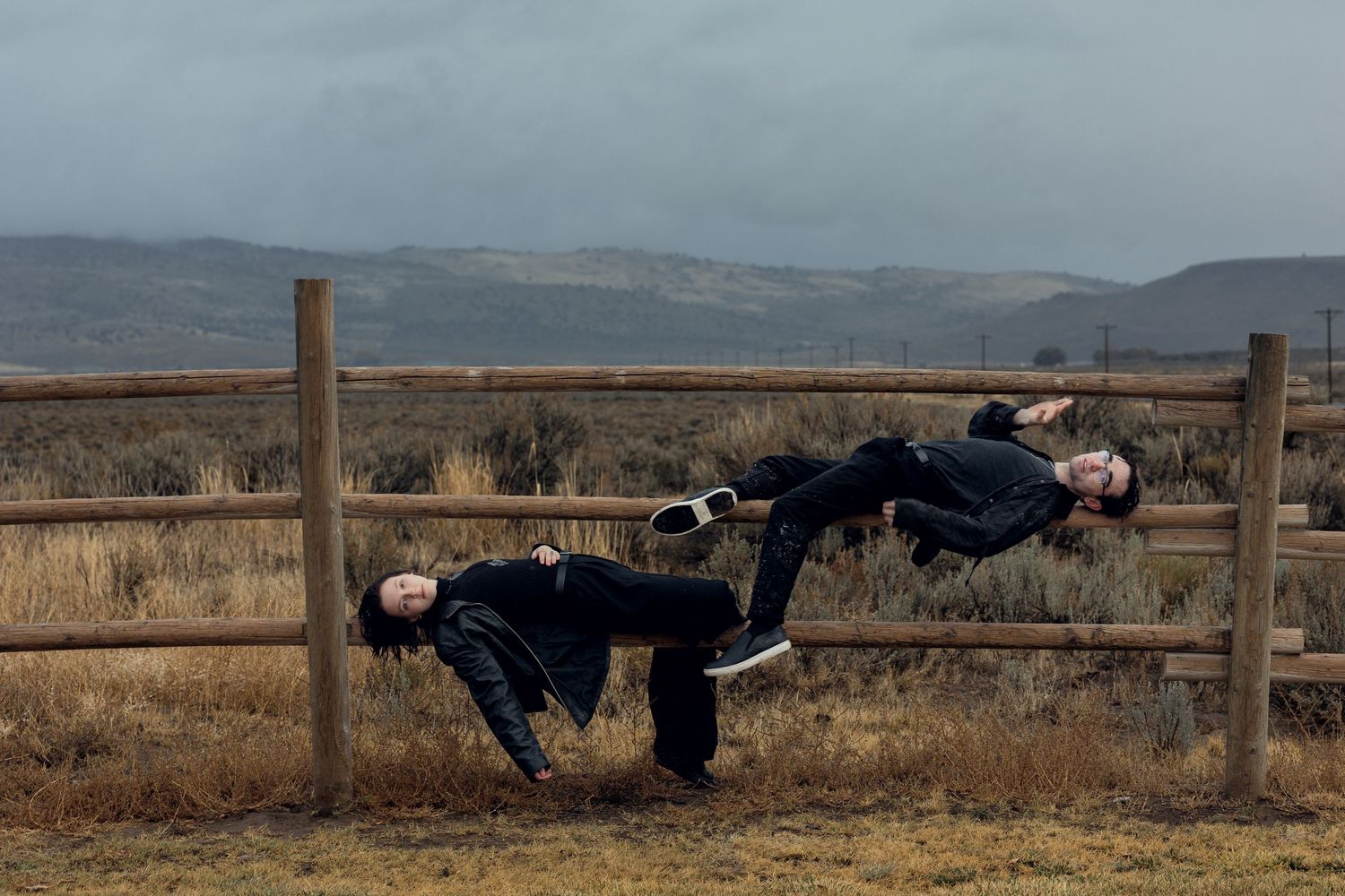 Two people in black clothing appear to levitate horizontally beside a rustic wooden fence in an open landscape.
