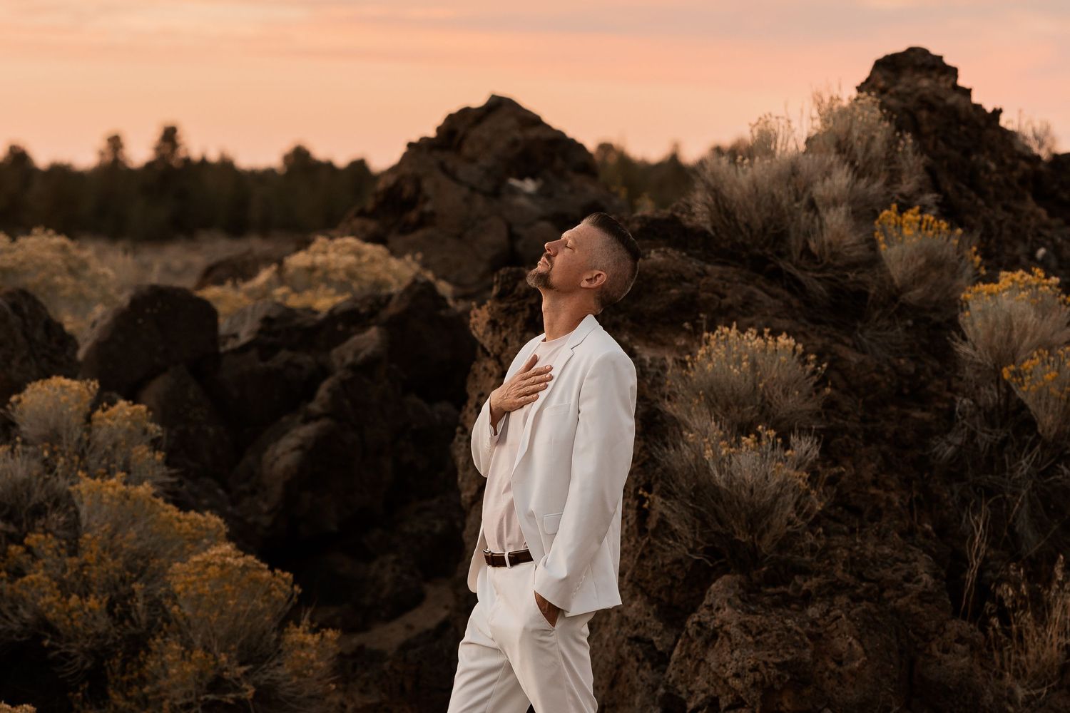 Man in white suit standing among dark rocks and sagebrush at sunset with head tilted back.