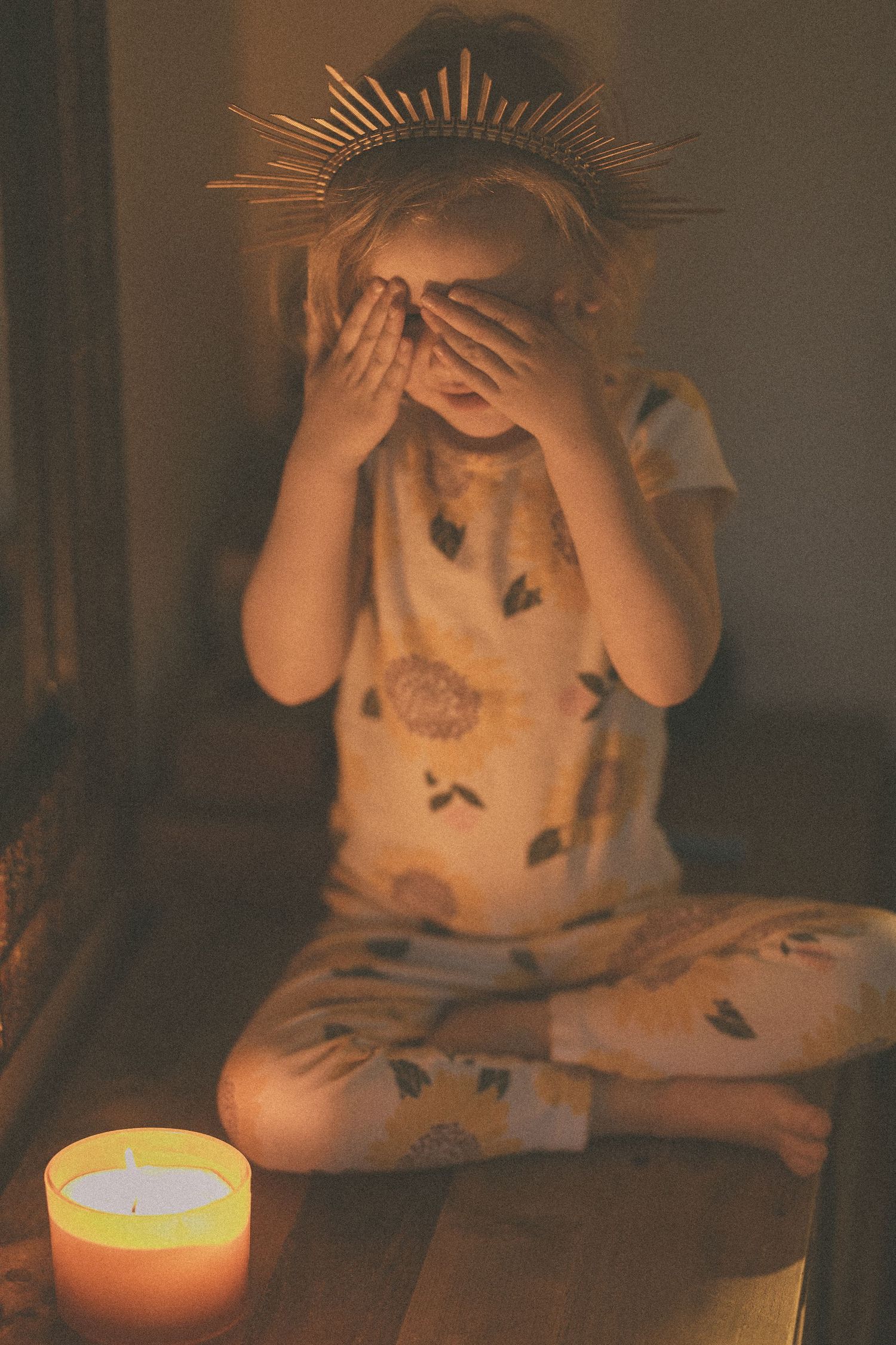 Child wearing a gold crown sits cross-legged covering eyes with hands beside a lit candle in dim light.