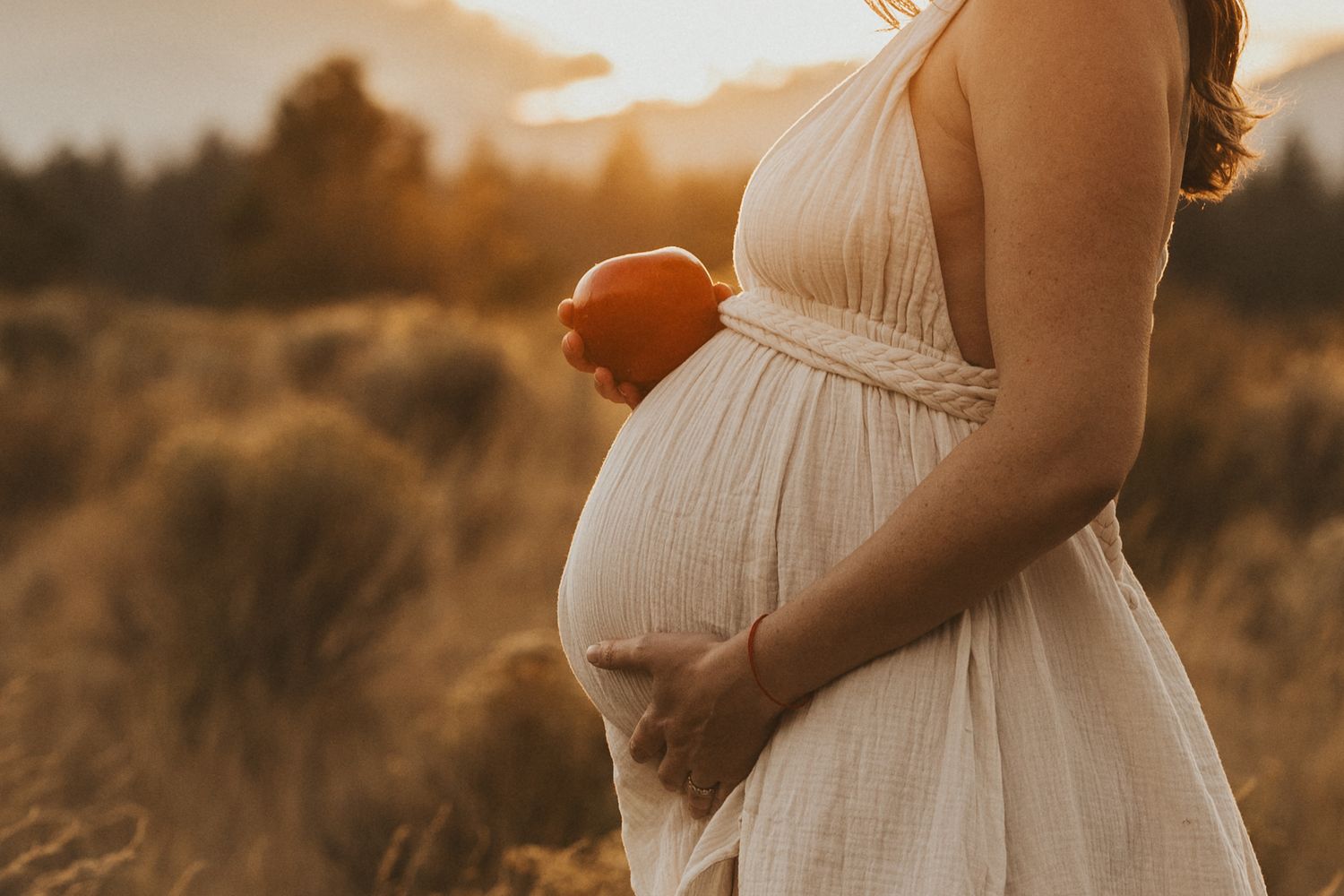 Pregnant woman in cream halter dress holding an apple beside her baby bump at golden sunset.