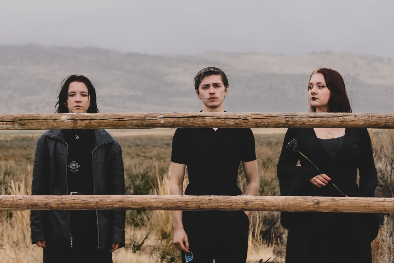 Three young adults dressed in black stand solemnly before a rustic wooden fence in a foggy rural landscape.