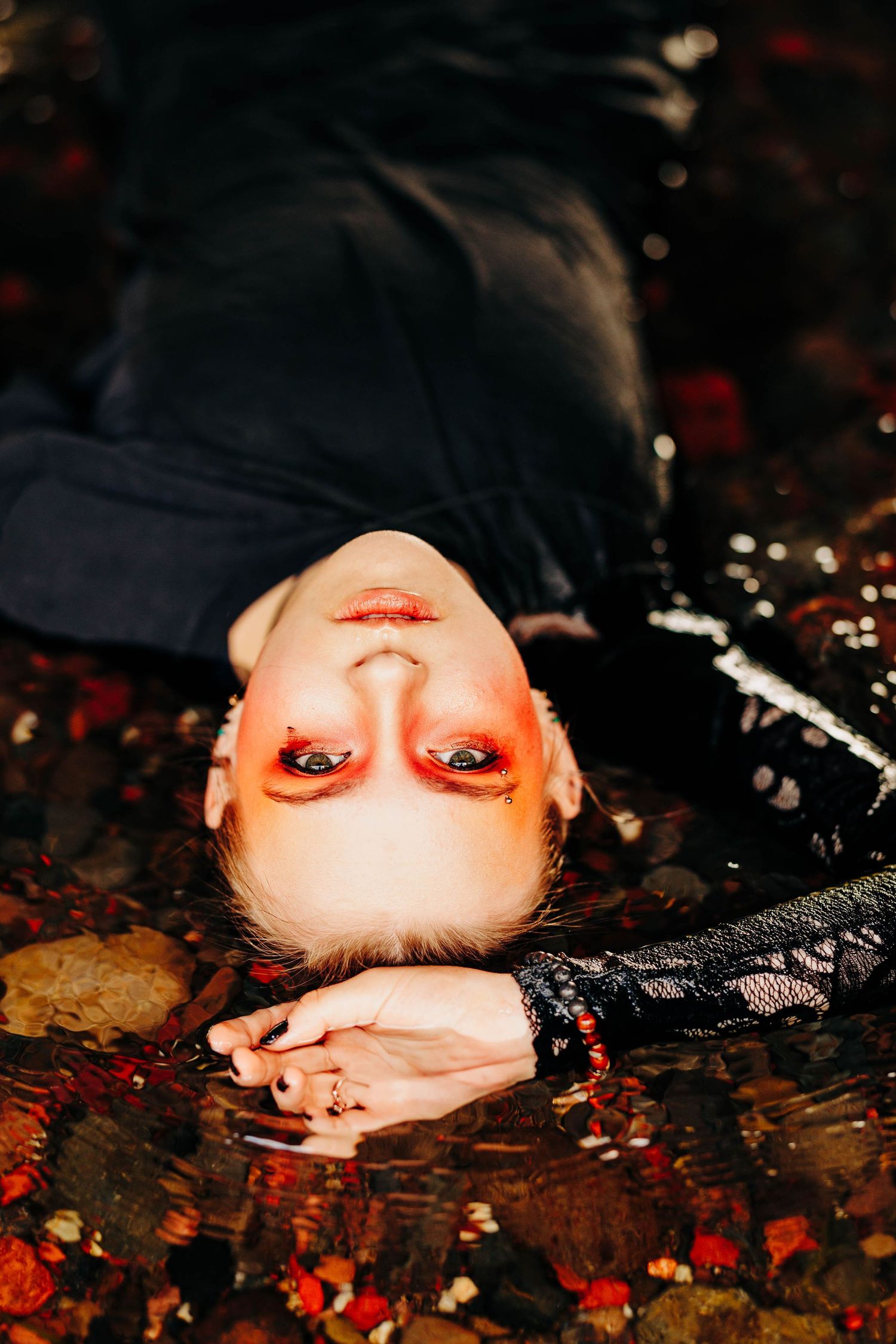 Woman with bold red makeup lies back in shallow rocky water wearing black lace sleeve.
