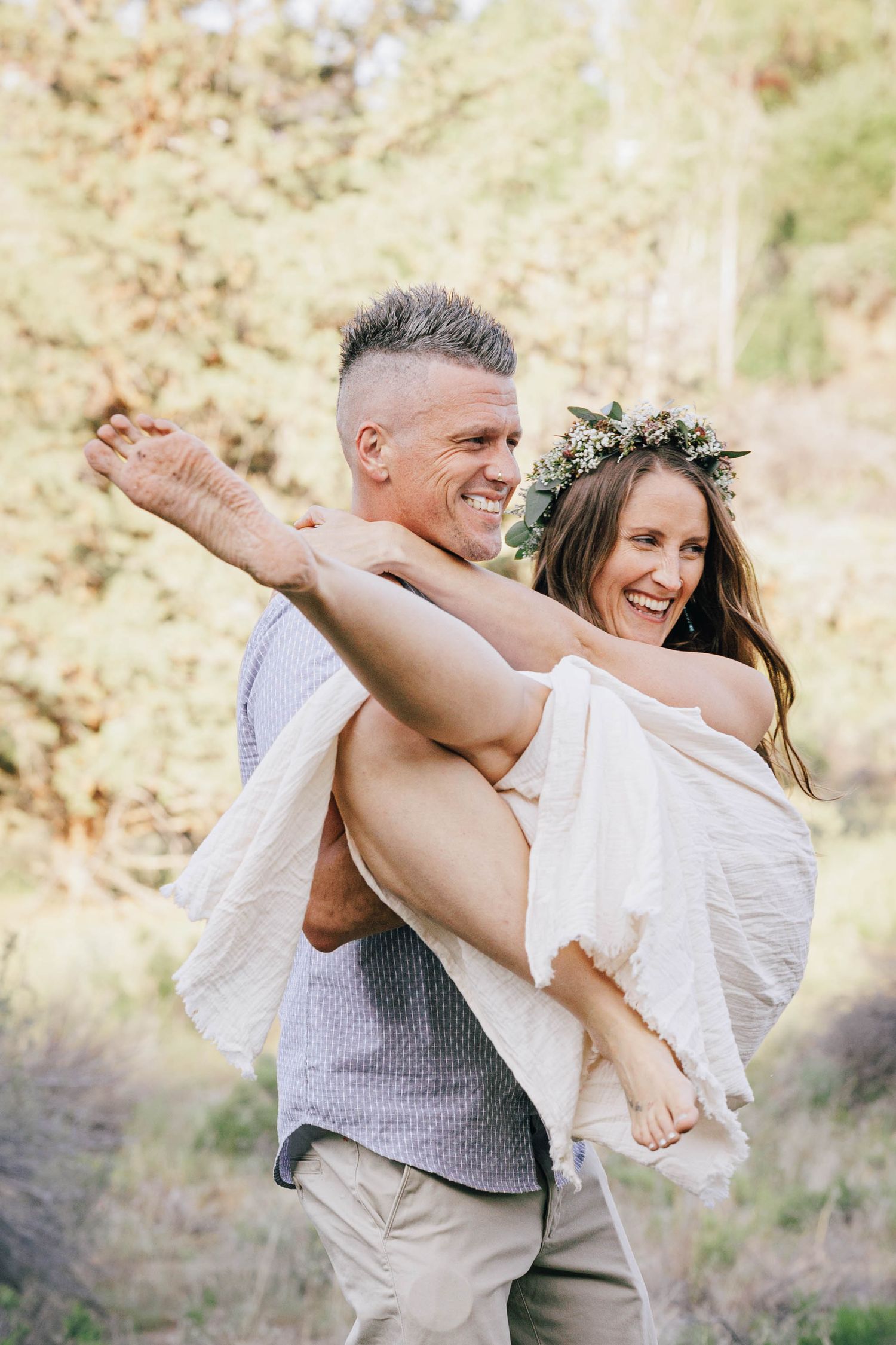 Man carrying laughing woman in white dress and floral crown outdoors.