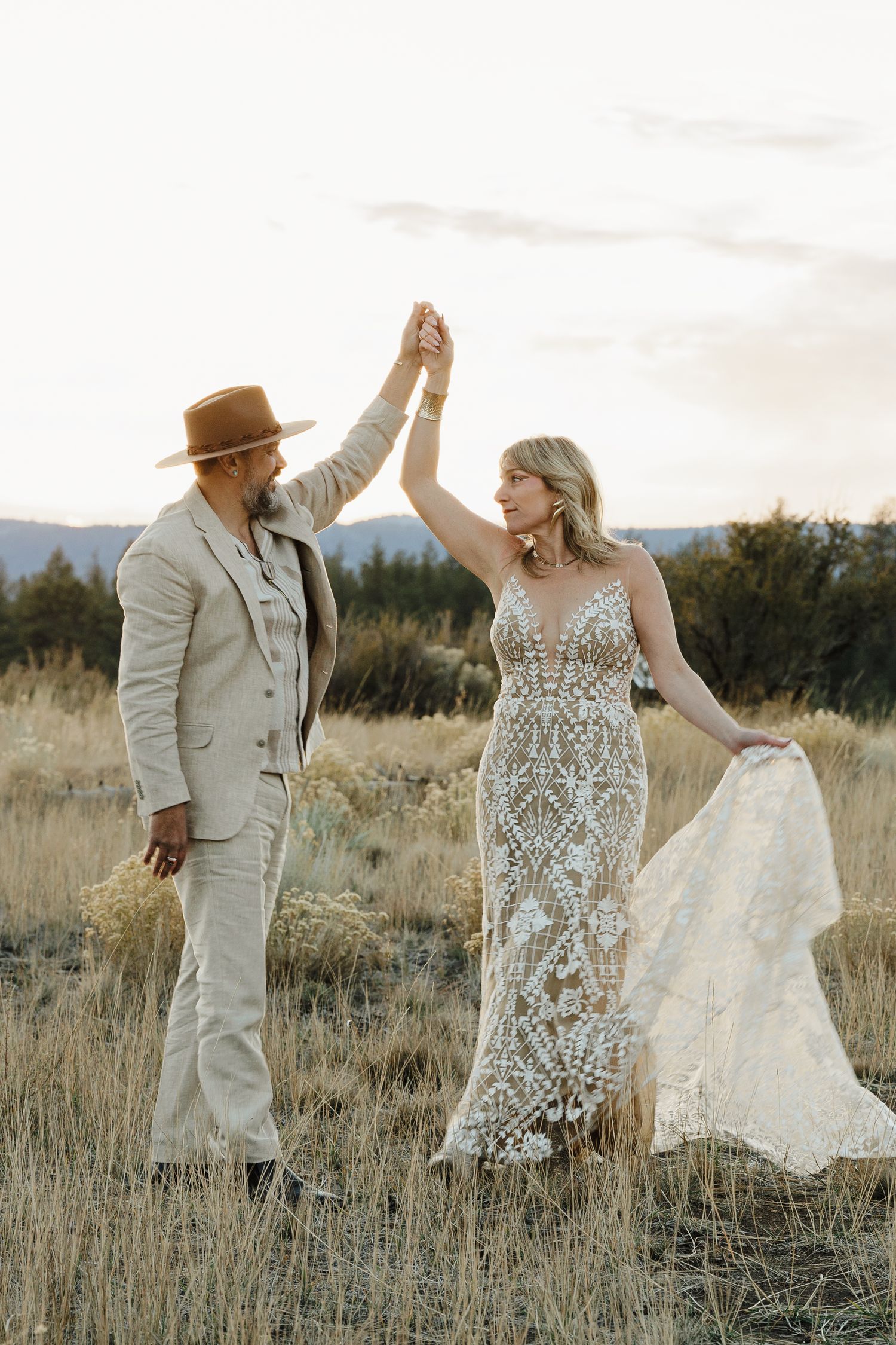 Bride and groom dance in golden meadow, she twirls in lace gown as mountains loom behind.