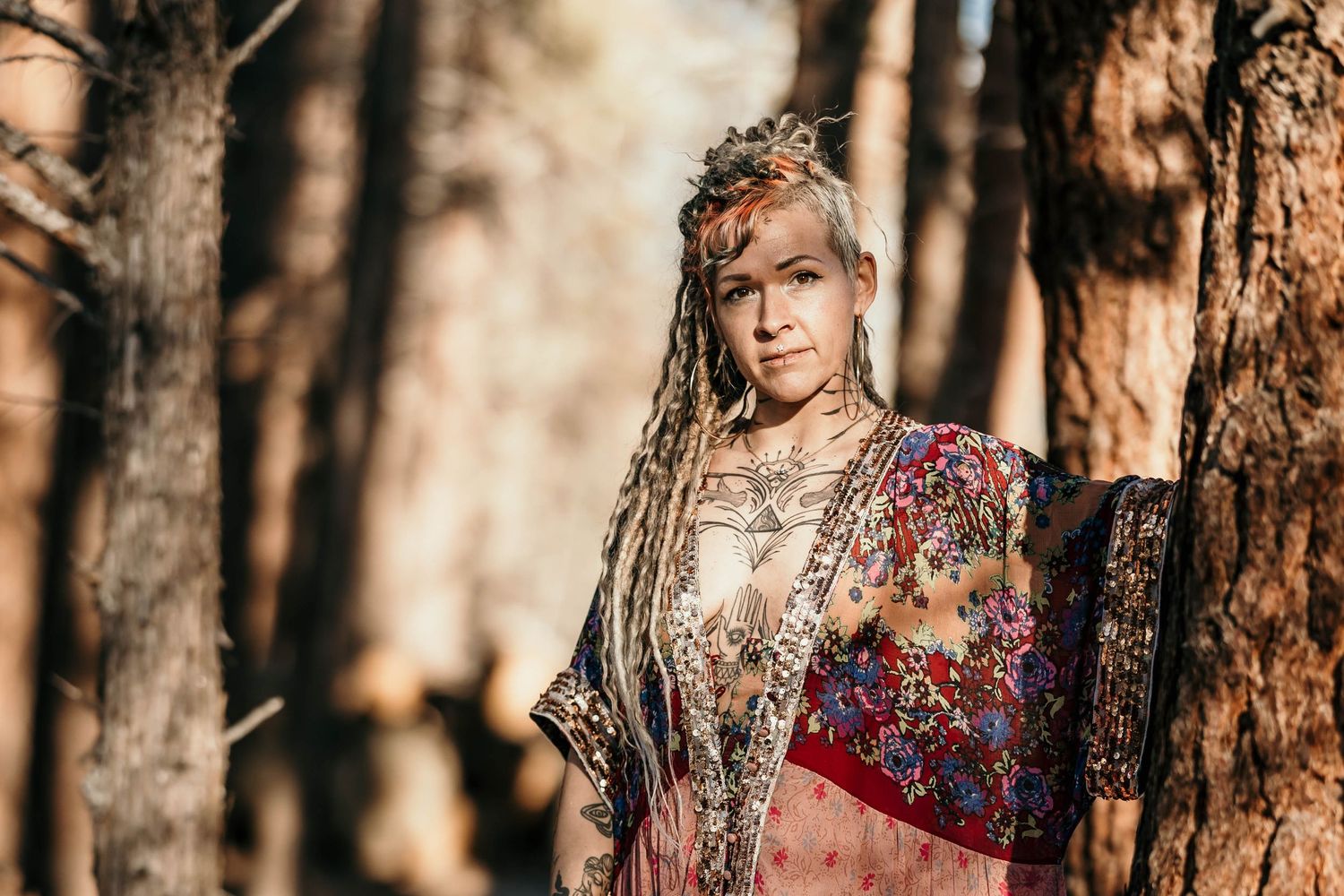 Tattooed woman with long dreadlocks and crown wearing a floral kimono standing among trees.