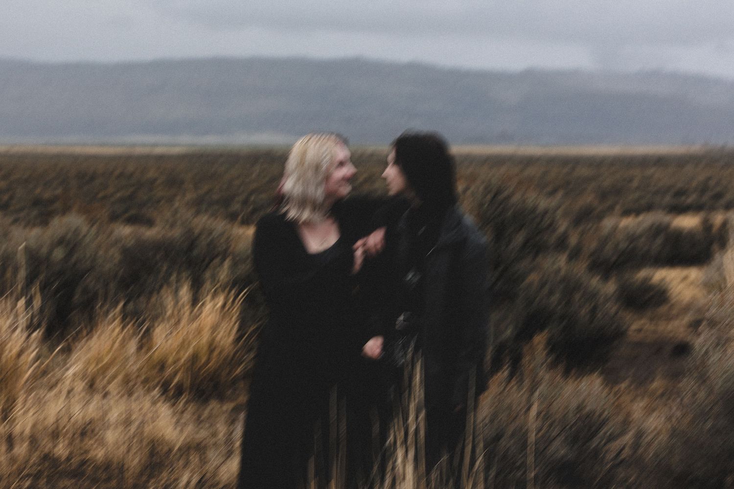 Two women in black clothing facing each other in a blurred artistic shot in a wild field.
