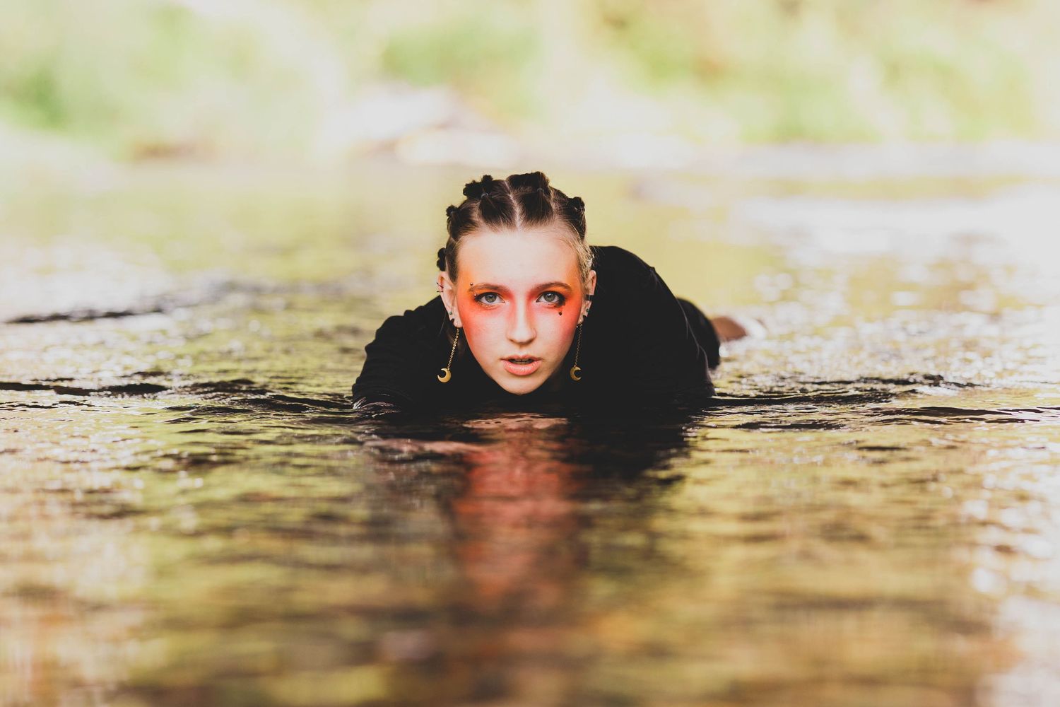 A young woman with red-tinted eye makeup and braided hair emerges from shallow water in a dramatic outdoor portrait.