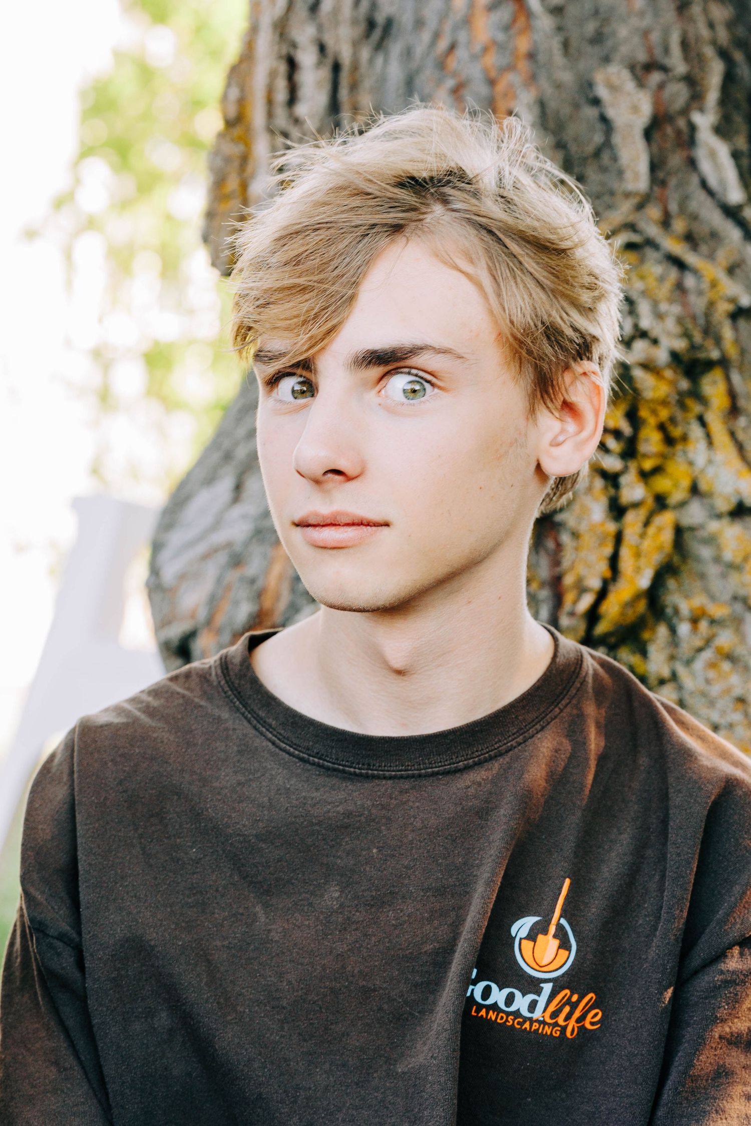Blonde teenage boy with green eyes in brown Good Life Landscaping tee by a tree.