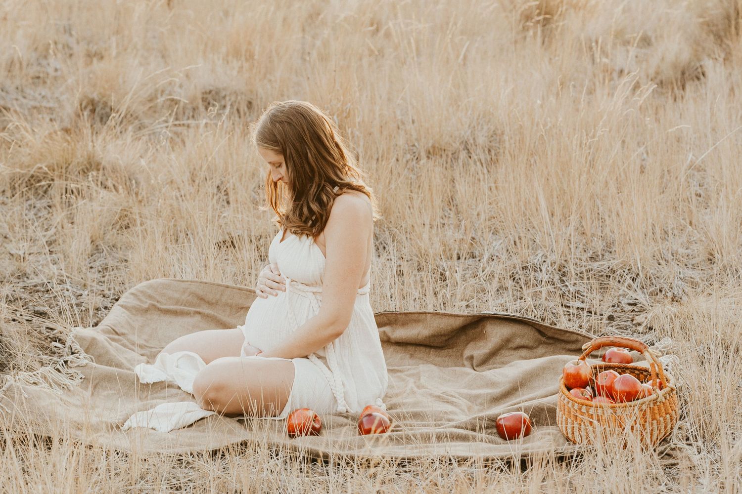 Pregnant woman in white dress sitting on blanket in dry grass field surrounded by fresh apples.