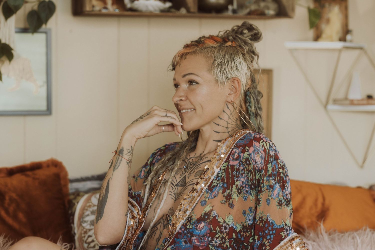 Smiling tattooed woman with dreadlocks wearing a floral bohemian kimono sitting on a couch.