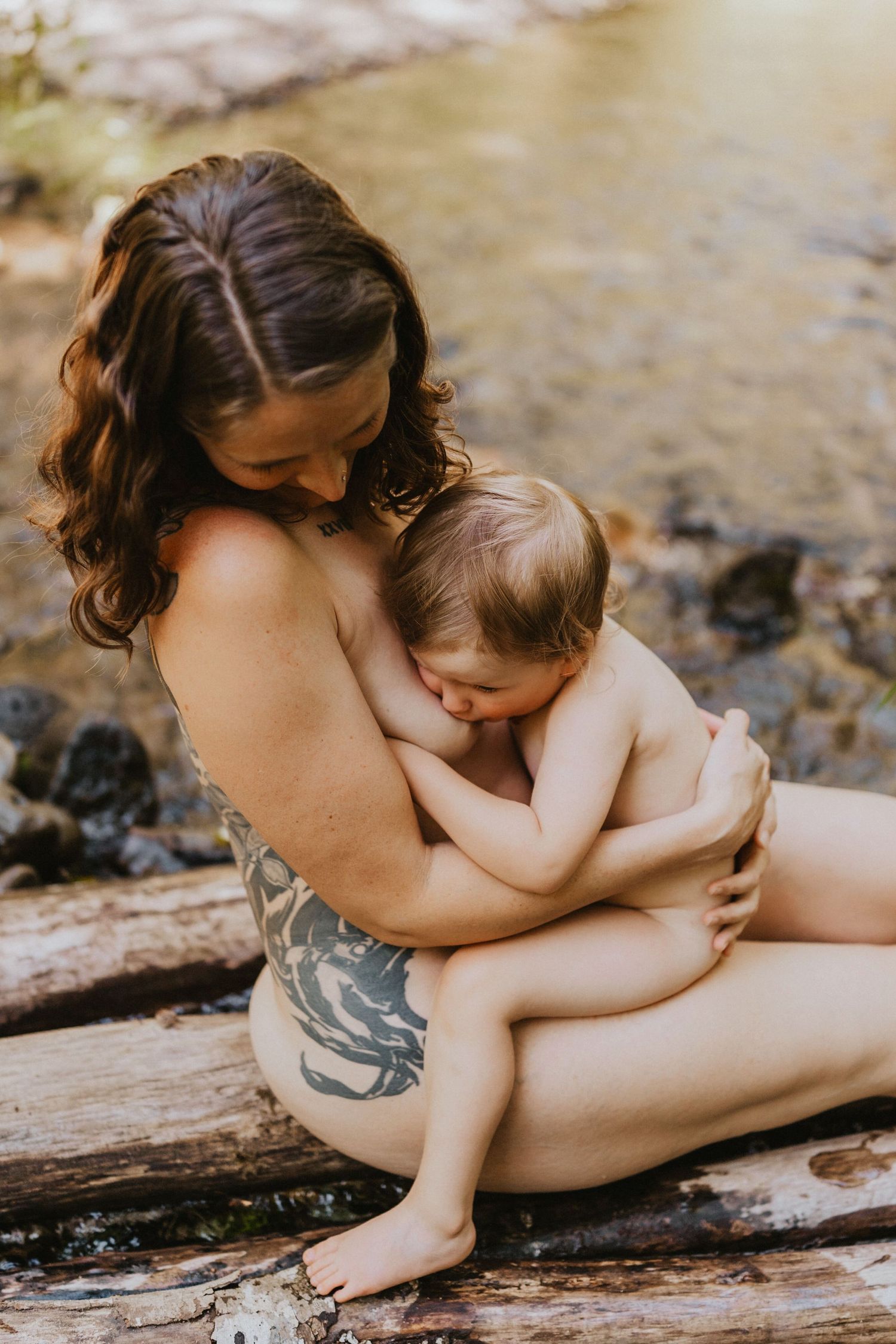 Mother breastfeeding baby while sitting on rocks near a stream in a natural outdoor setting.