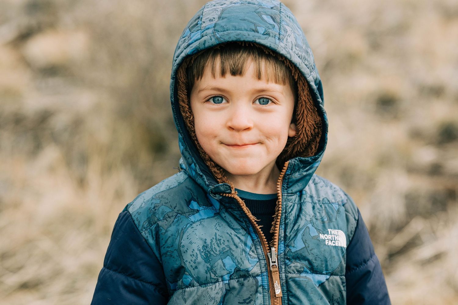 Smiling young boy wearing a blue hooded North Face puffer jacket outdoors.
