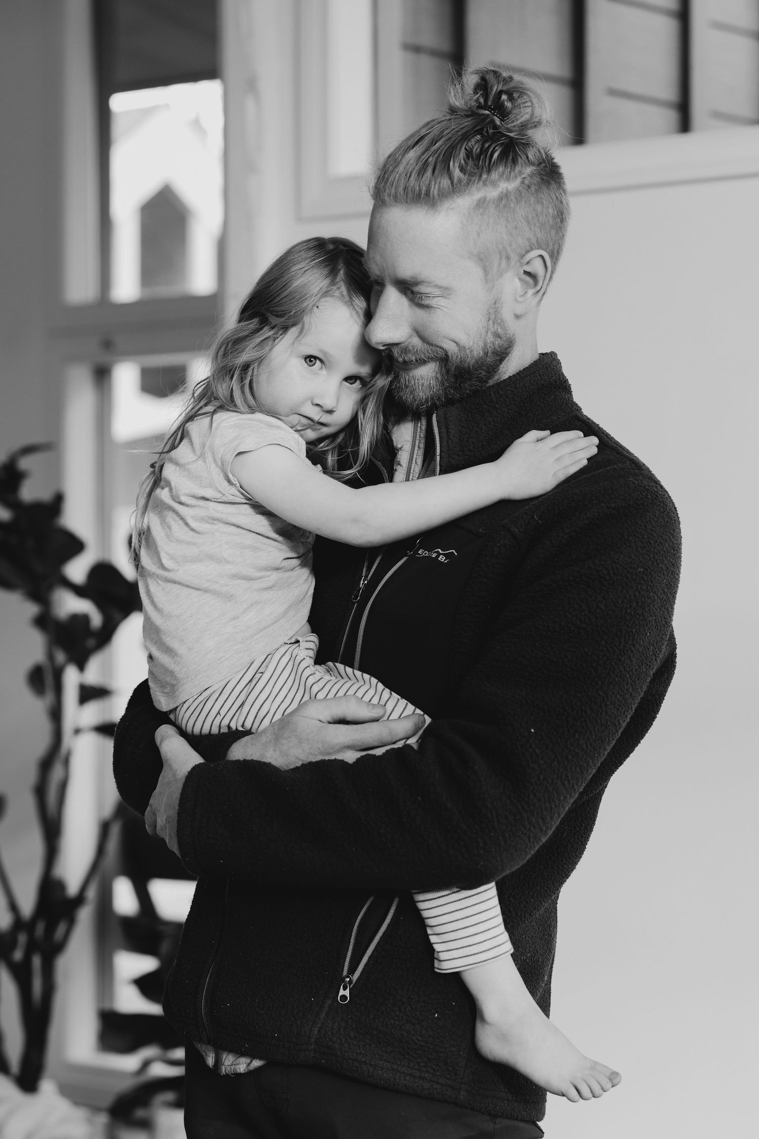 Black and white photo of bearded father with man bun lovingly holding young daughter outdoors.