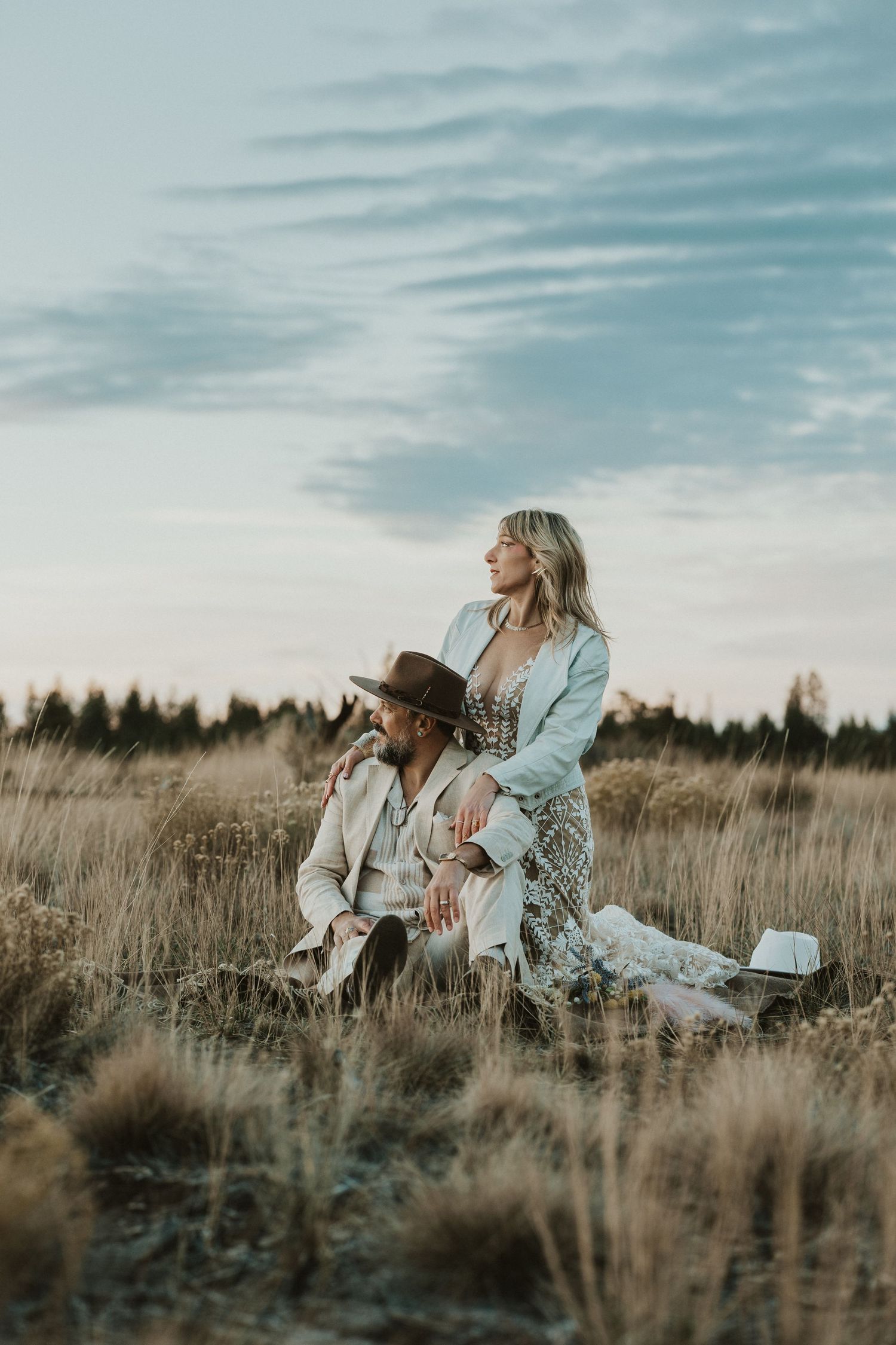 Elegant boho couple sitting in a golden meadow at dusk with pine forest backdrop.