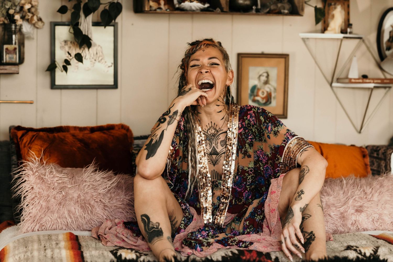 Tattooed woman laughs joyfully while sitting on couch in bohemian styled interior.