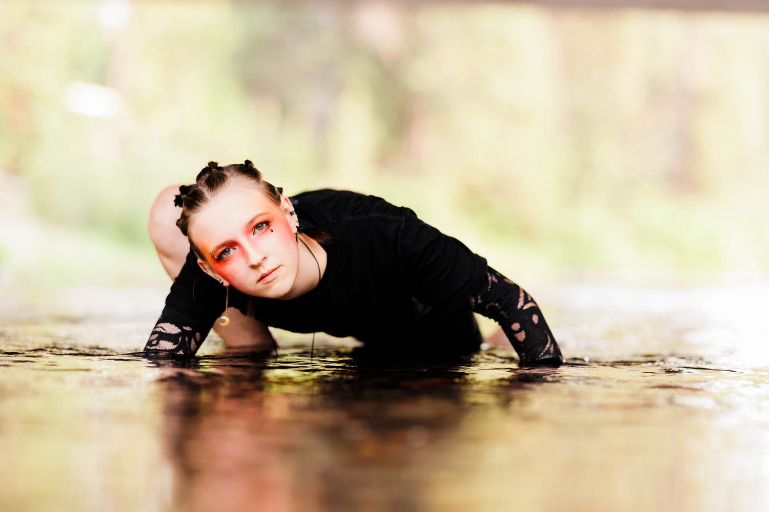 A young woman with red eye makeup crawls through shallow water wearing a black outfit in a nature setting.