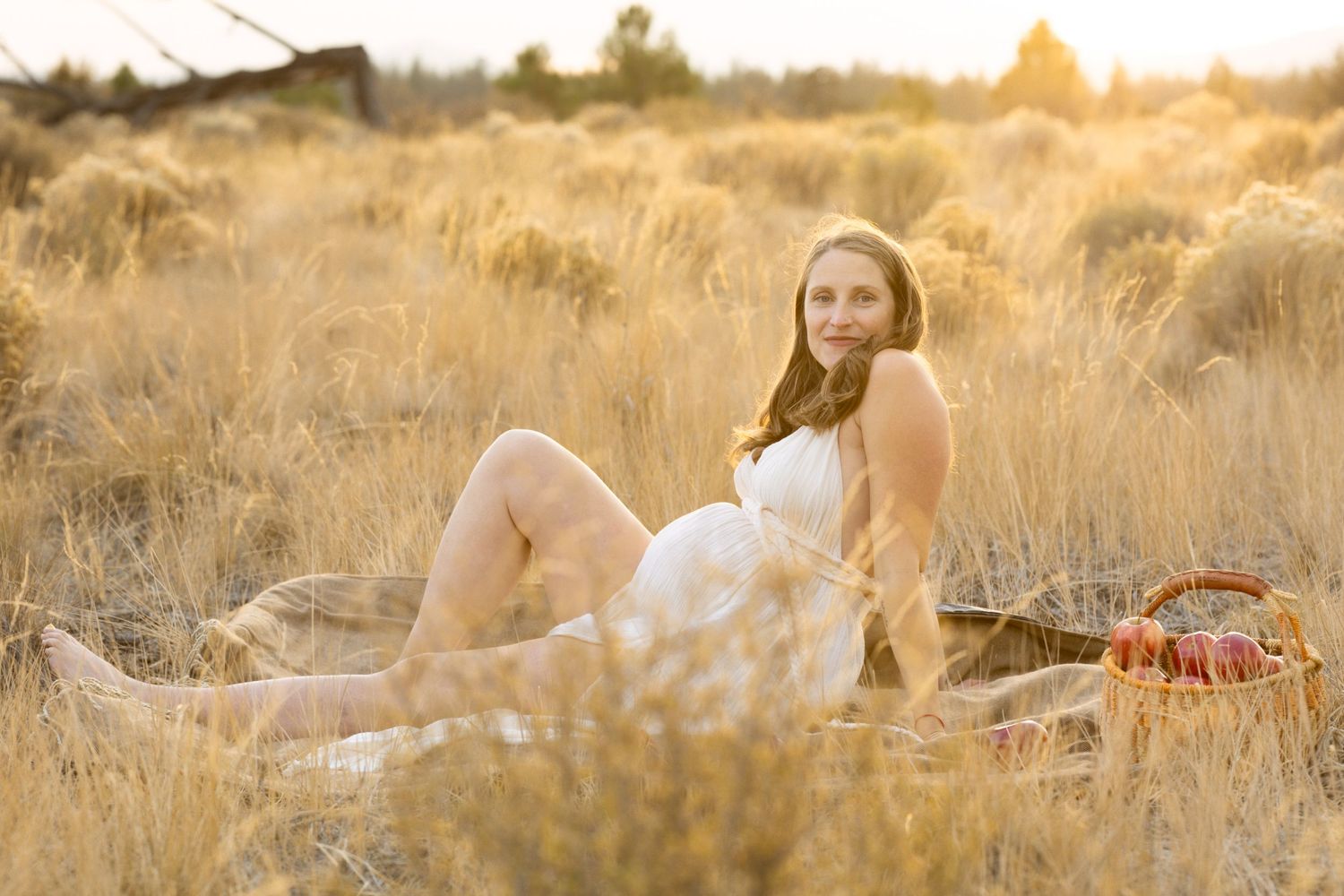 Smiling pregnant woman in white dress sitting in sunlit golden field with a wicker basket nearby.