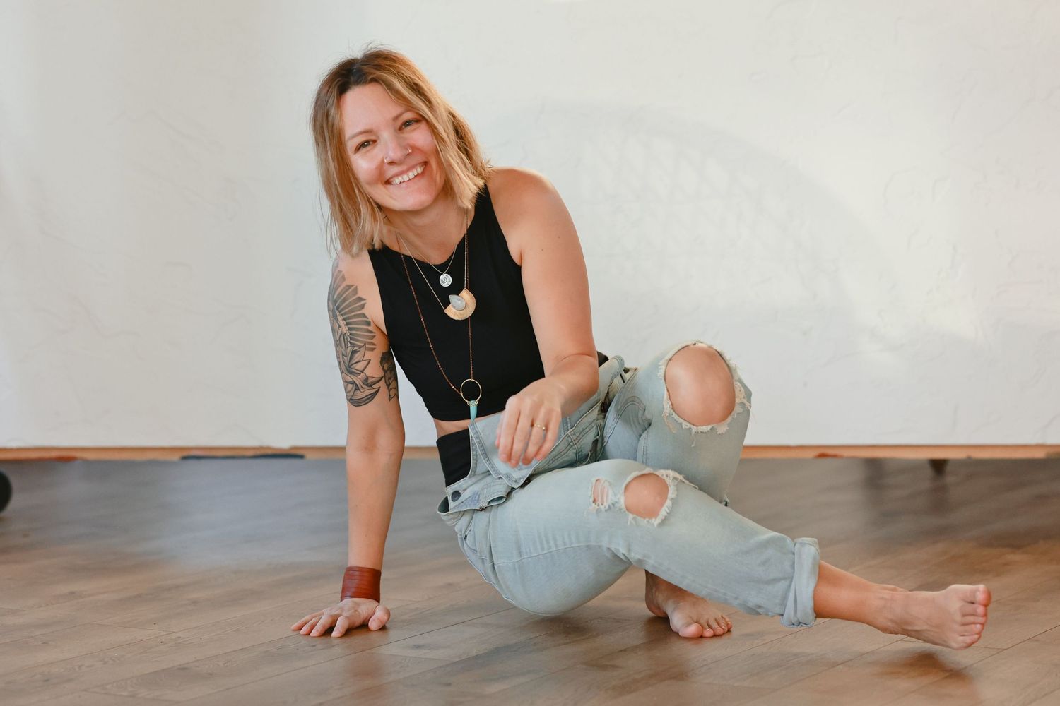 Smiling woman with tattoos sits on wooden floor in ripped jeans and black tank top, wearing layered necklaces.