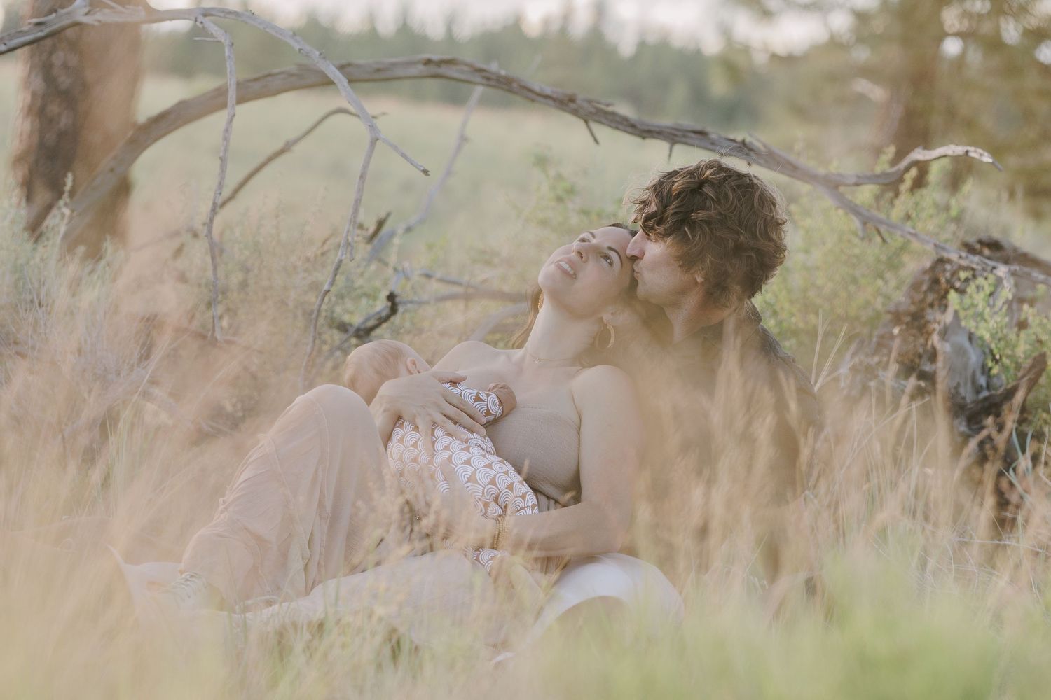 Couple sitting intimately in tall golden grass with baby, surrounded by natural outdoor wilderness scenery.