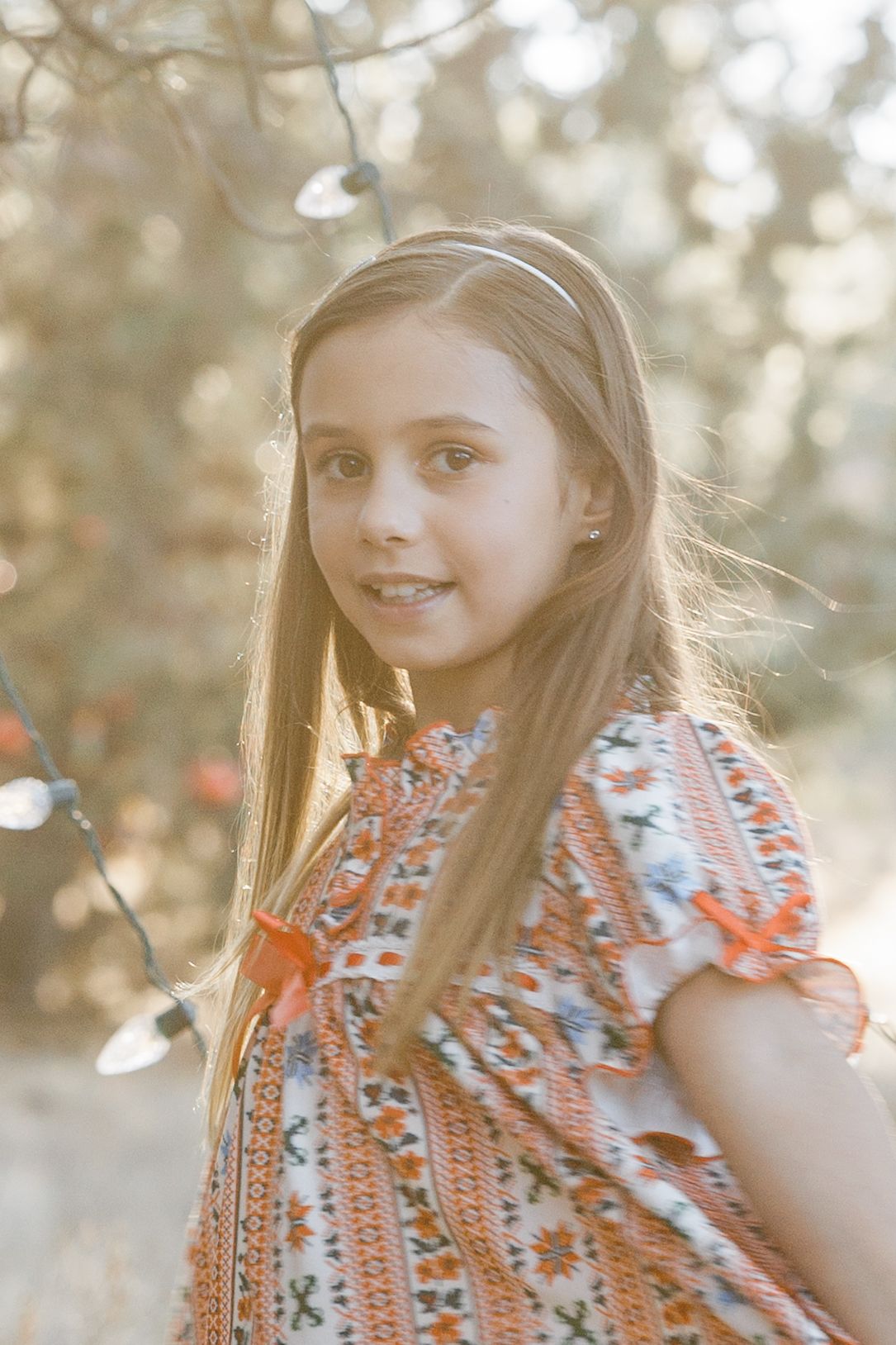 Pretty girl with long brown hair wearing patterned dress holds string lights in sun-dappled outdoor setting.