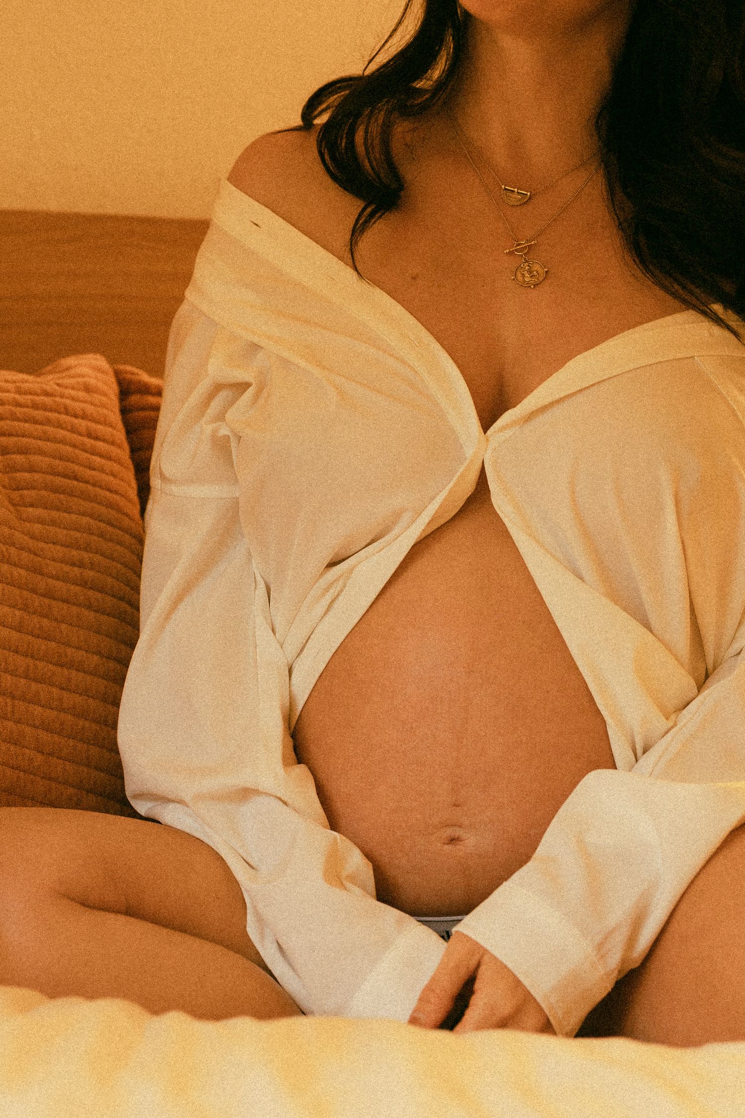 Pregnant woman in open white button-up shirt cradling bare baby bump while sitting on bed with gold necklace.