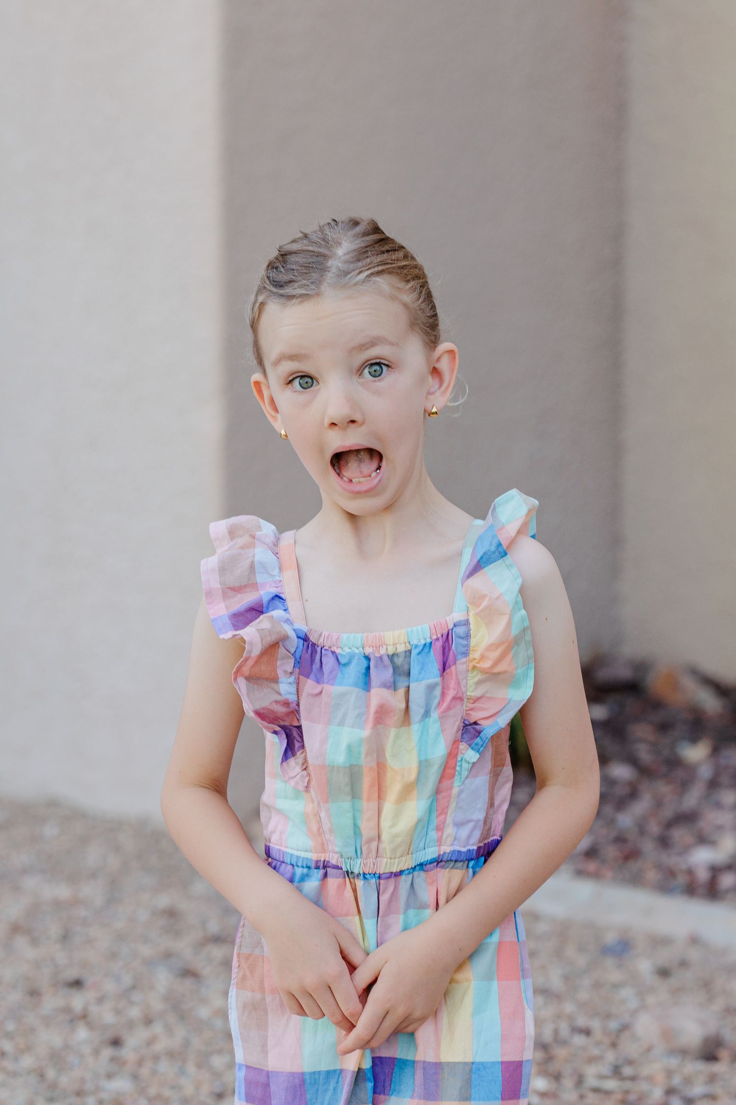 Surprised young girl in colorful pastel plaid romper makes funny face with mouth open outdoors.