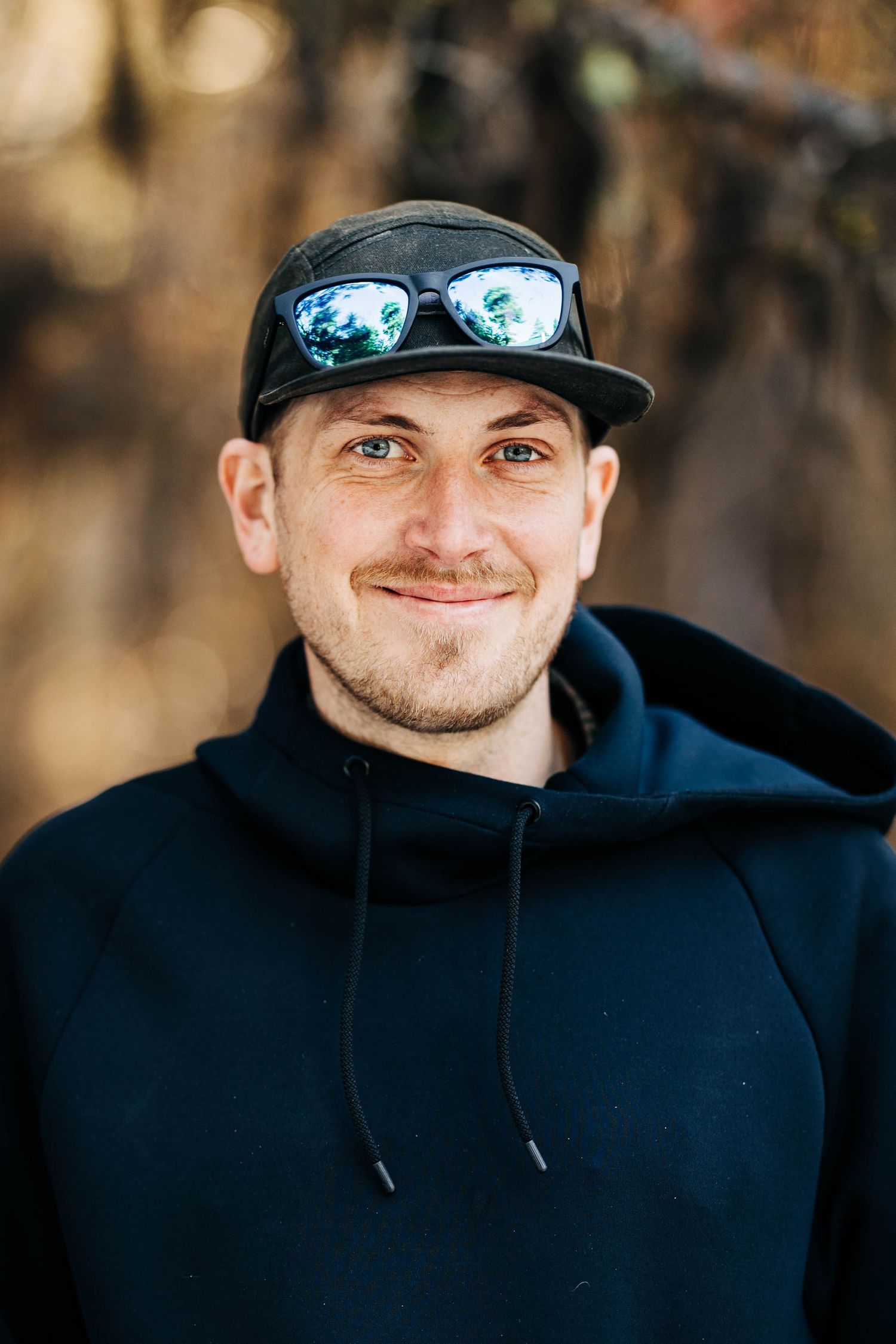 Smiling man in black hoodie and cap with sunglasses outdoors.