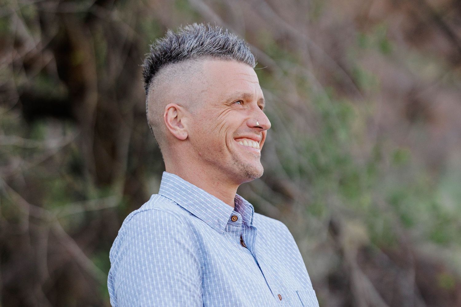 Smiling middle-aged man with short gray hair wearing a light blue shirt outdoors in natural setting.