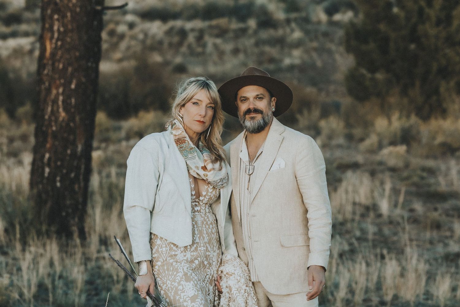 Stylish boho couple posing together in a sun-drenched meadow with pine trees in background.