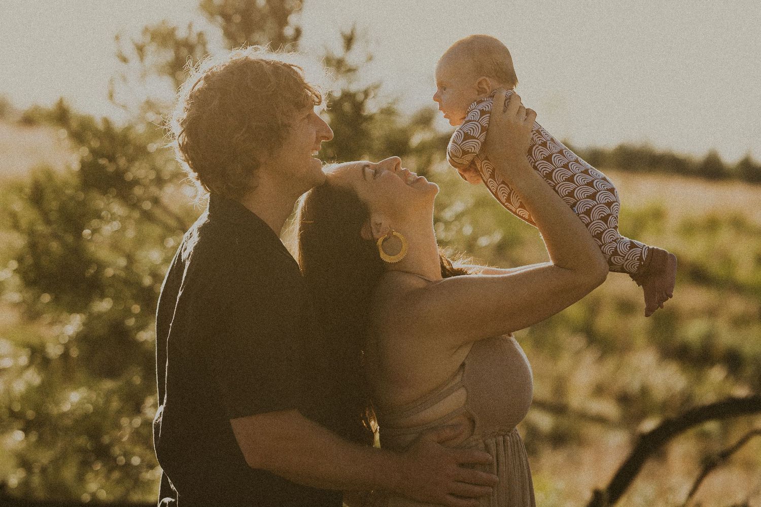 Happy parents lifting their newborn baby in the air during a golden hour outdoor photo session.
