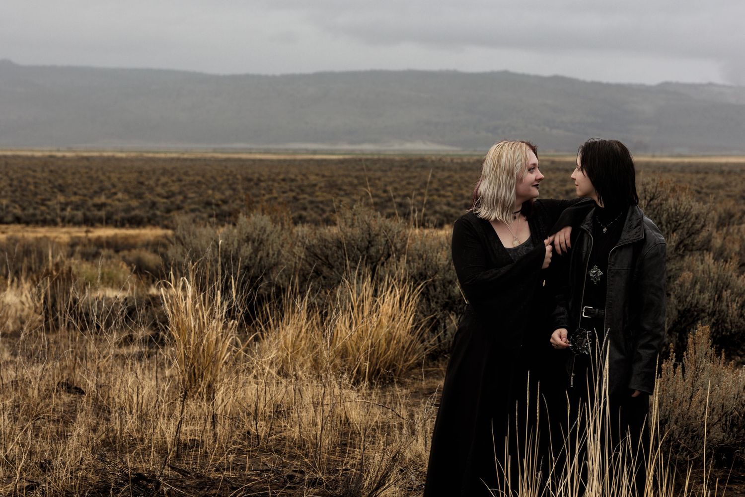 Two women in black dresses facing each other in a vast open desert field under cloudy skies.