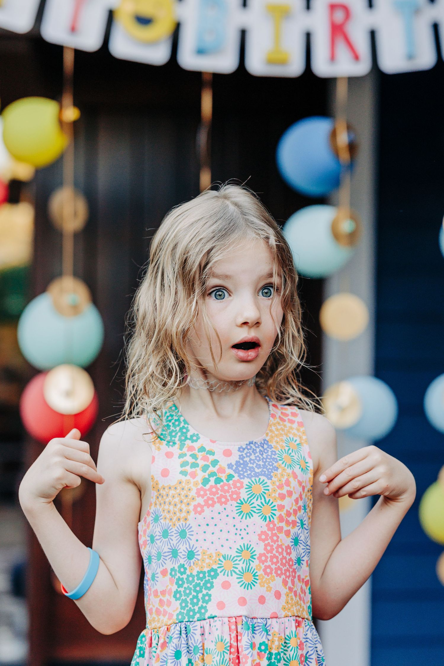 Astonished young girl in floral sundress points to herself at festive birthday party with balloon backdrop.