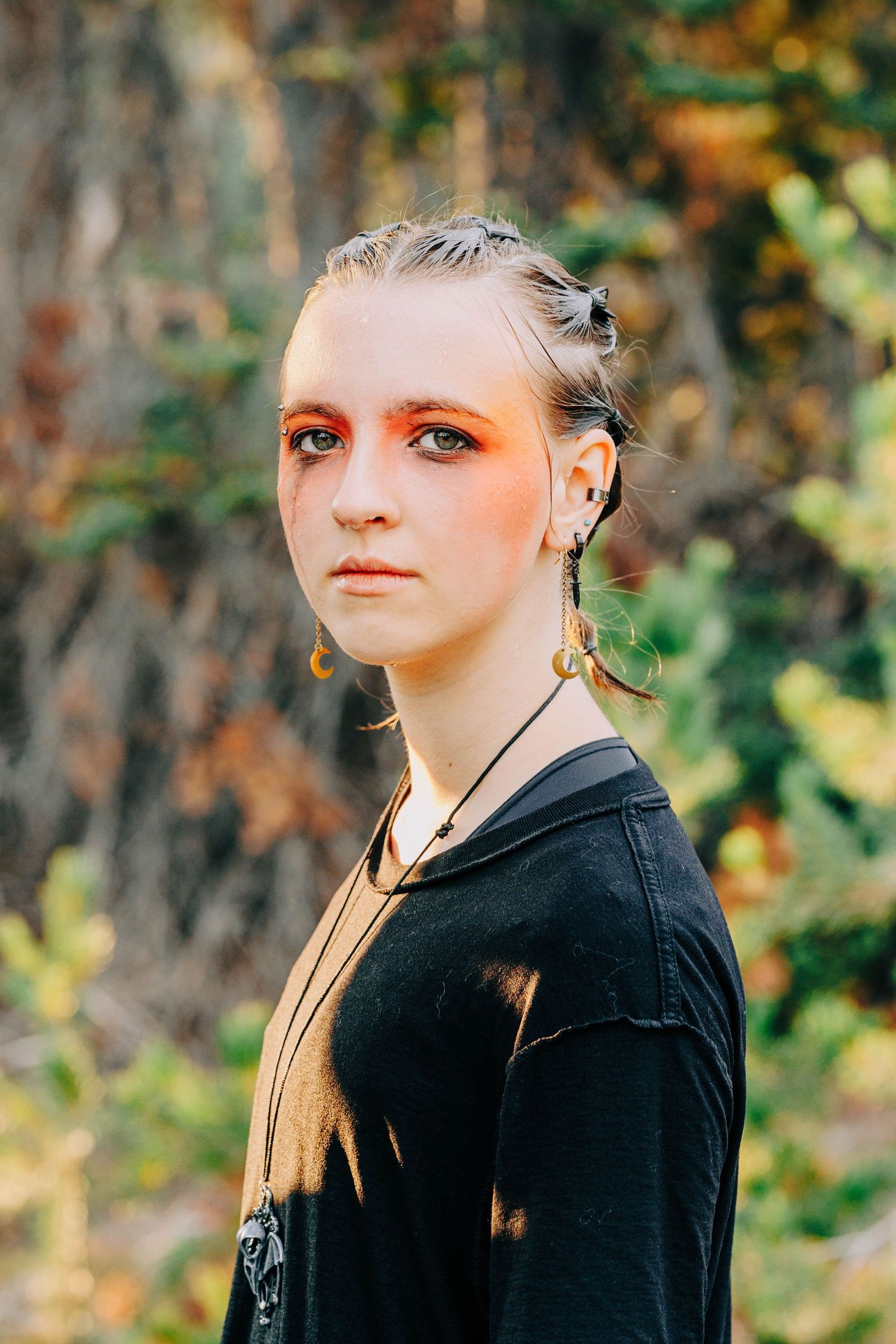 Young woman with orange eye makeup and dangling earrings poses outdoors in black hoodie against autumn foliage backdrop.