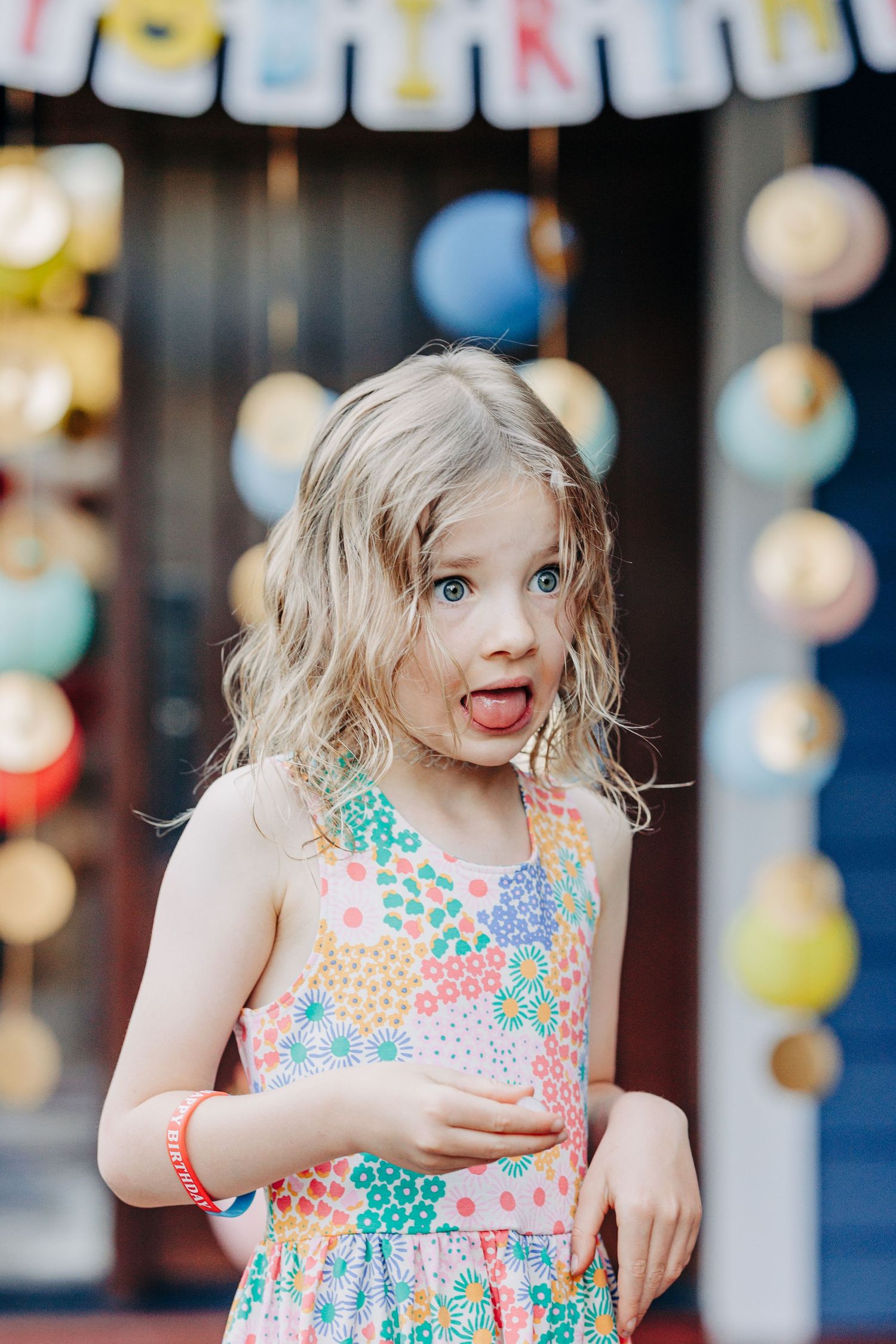 Playful young girl sticking out her tongue wearing a colorful floral sundress at a party.