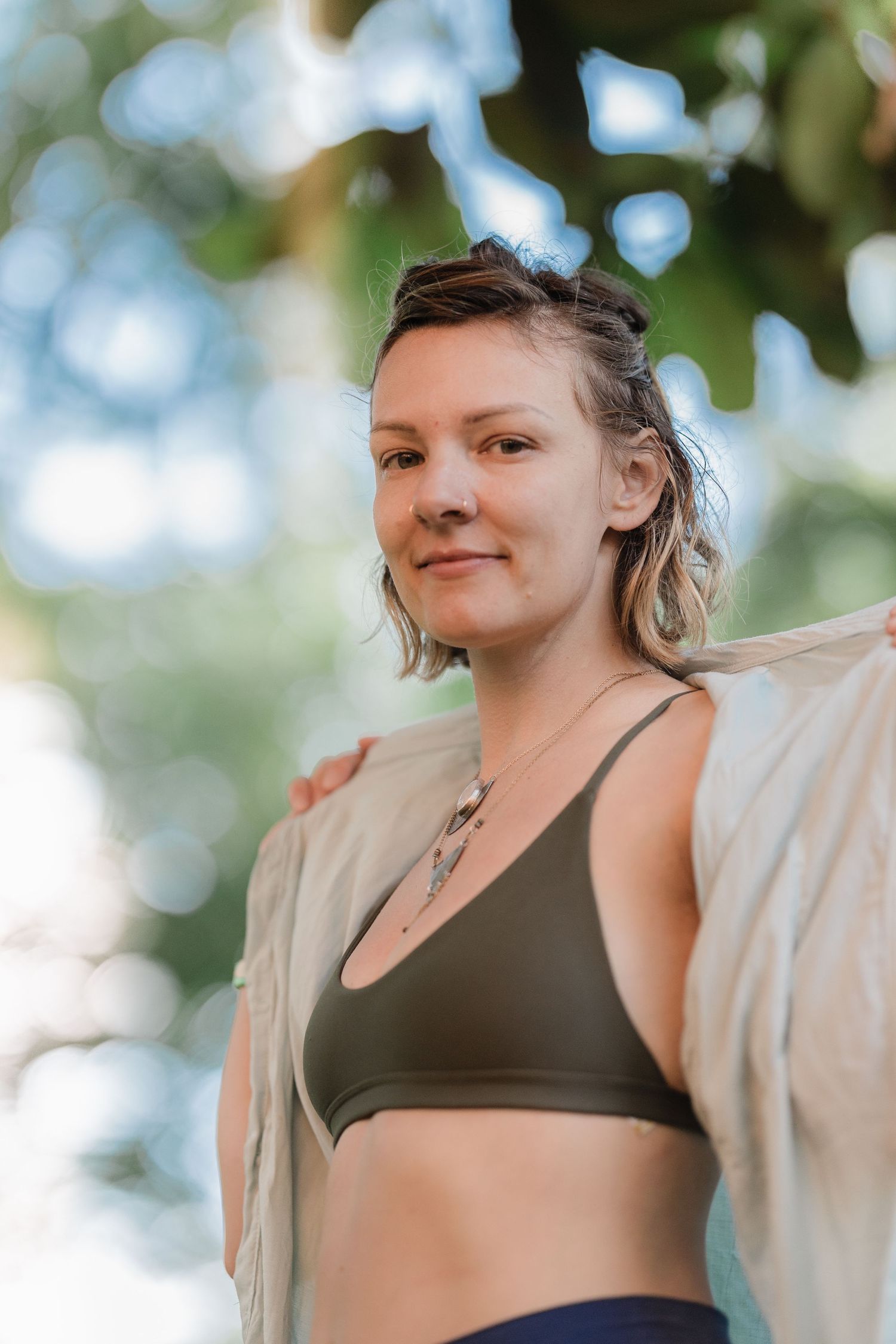 Young woman in an olive green sports bra smiling outdoors under lush green trees.