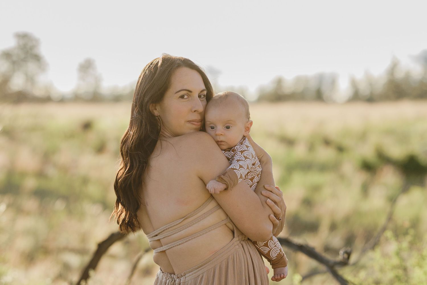 Mother in off-shoulder beige dress holding newborn baby in open field during golden hour photography.