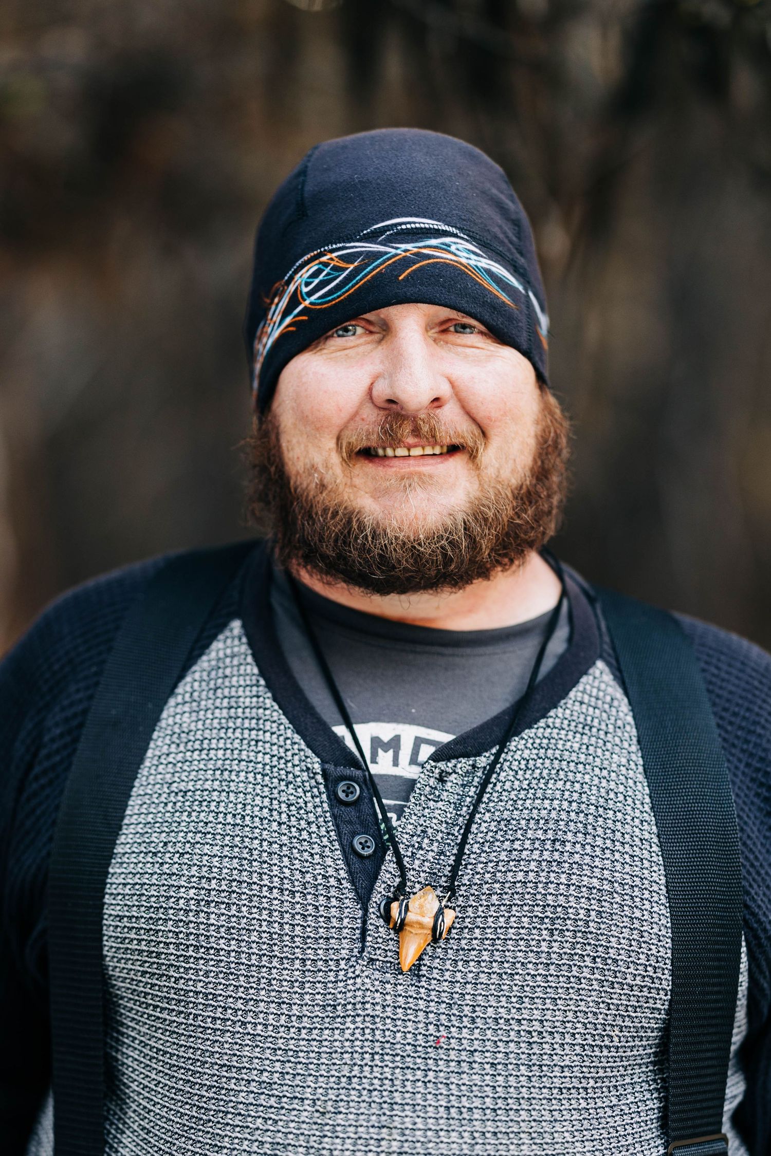 Bearded man in beanie and gray sweater smiling outdoors.