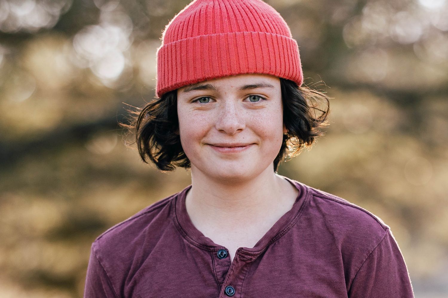 Smiling young person wearing a red beanie hat and purple henley shirt outdoors in natural light.
