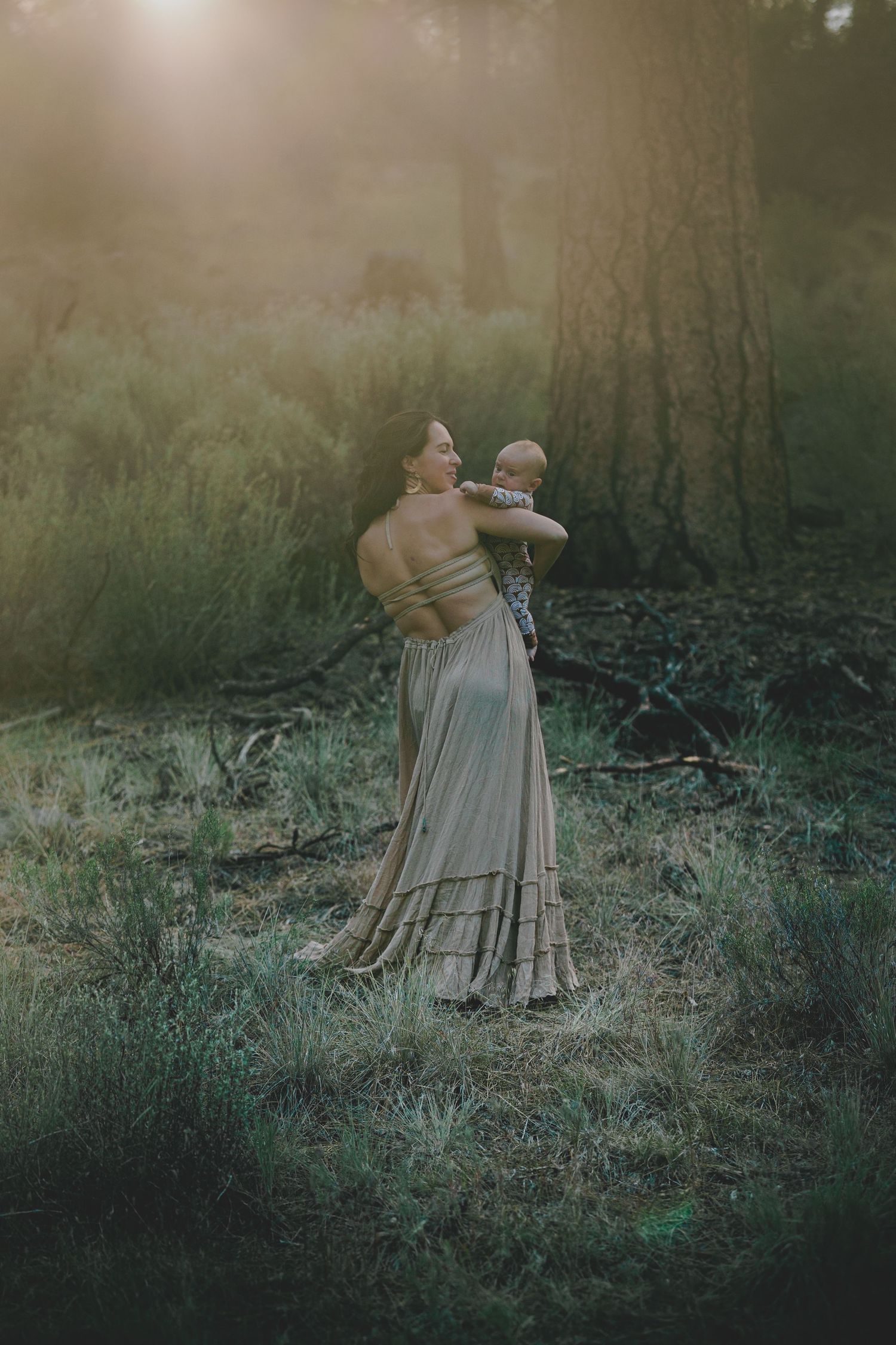 Mother in elegant gray gown holding newborn baby in sunlit forest during outdoor photo session.