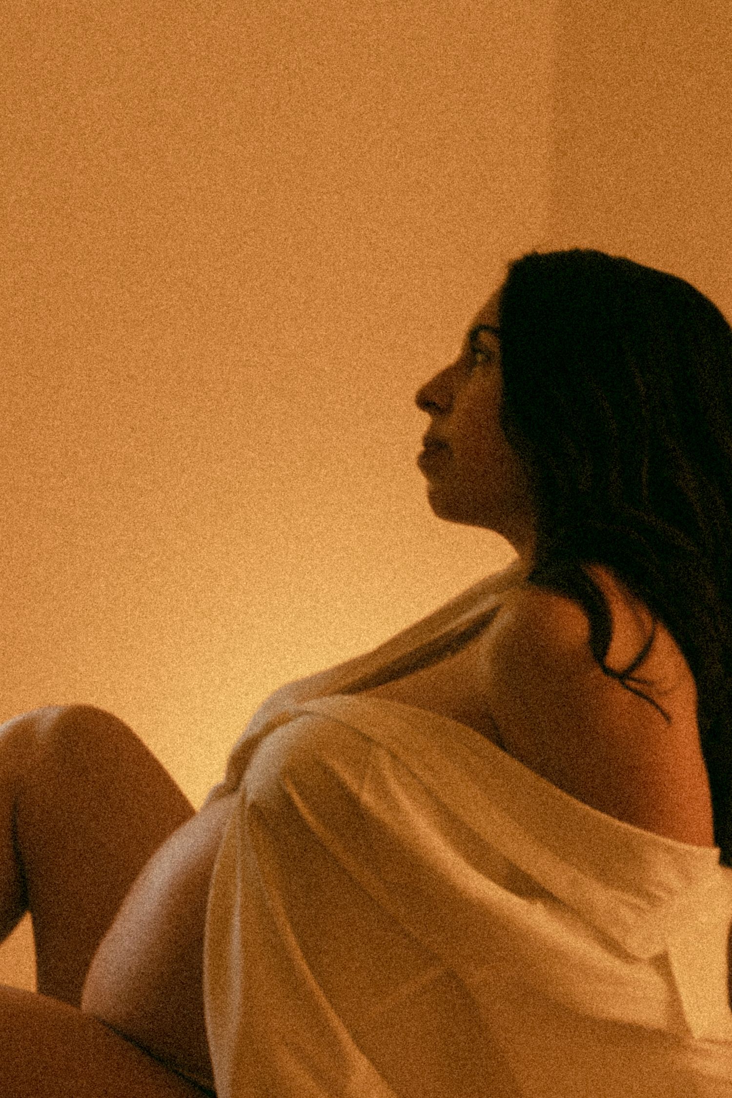 Pregnant woman wrapped in white fabric sitting with knees up gazing away in warm orange ambient light.