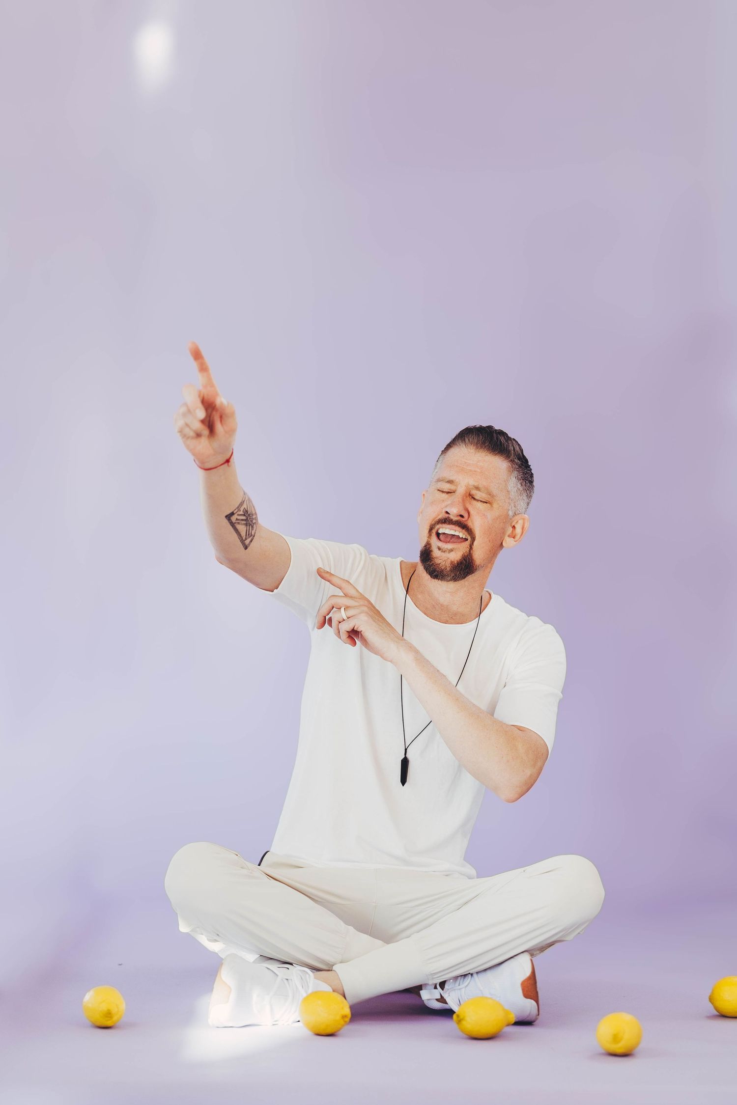 Tattooed man in white outfit sitting cross-legged on floor with lemons, pointing upward laughing.