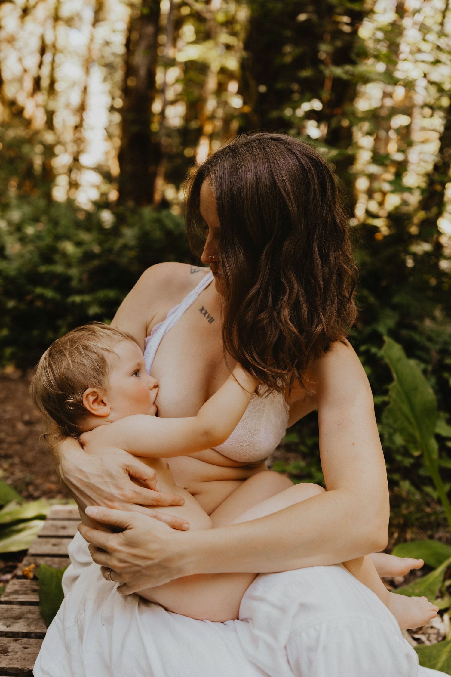 Mother breastfeeding her baby outdoors in a lush green forest setting, wearing a nude bra.