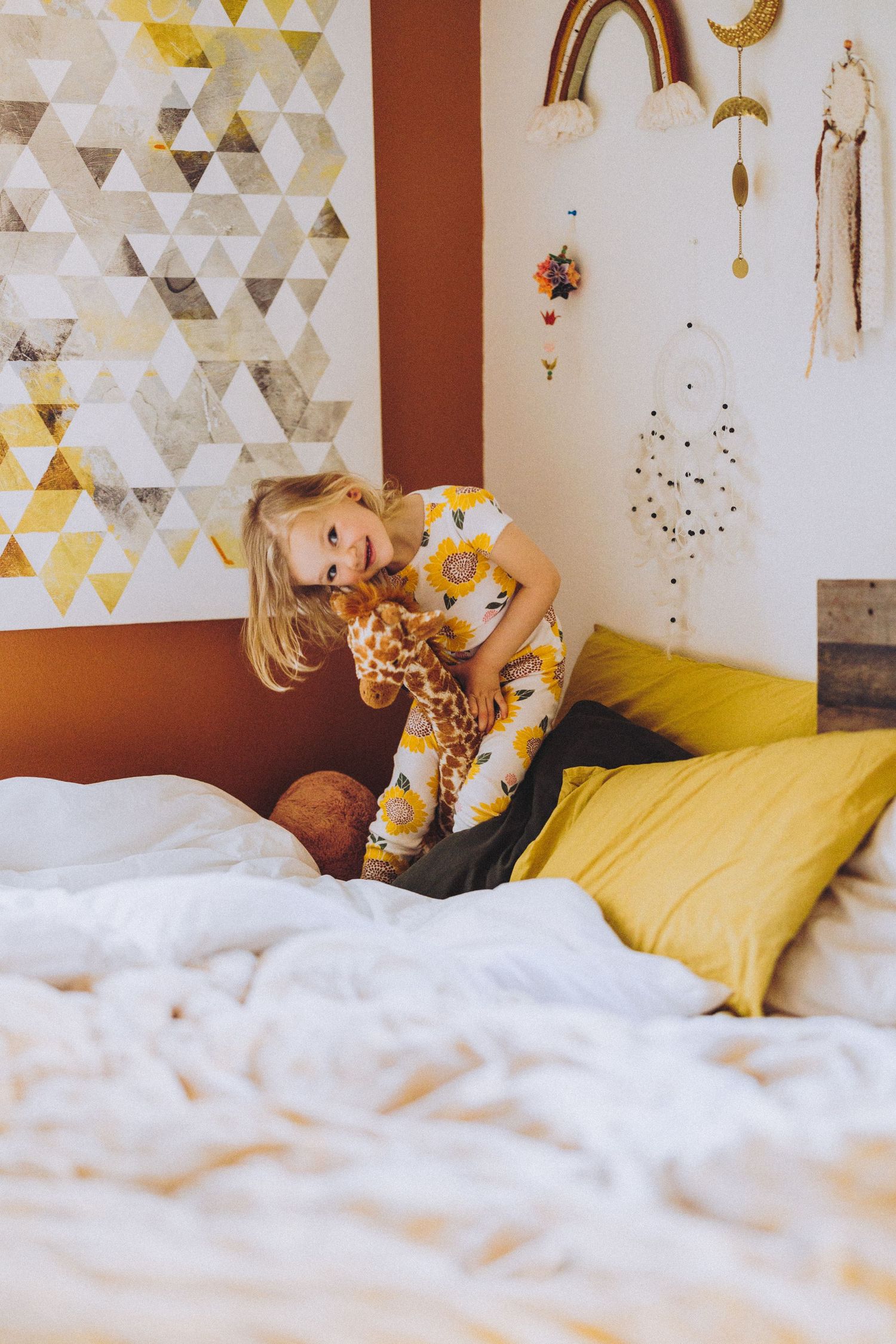Blonde girl in sunflower pajamas hugging giraffe stuffed animal on bed.