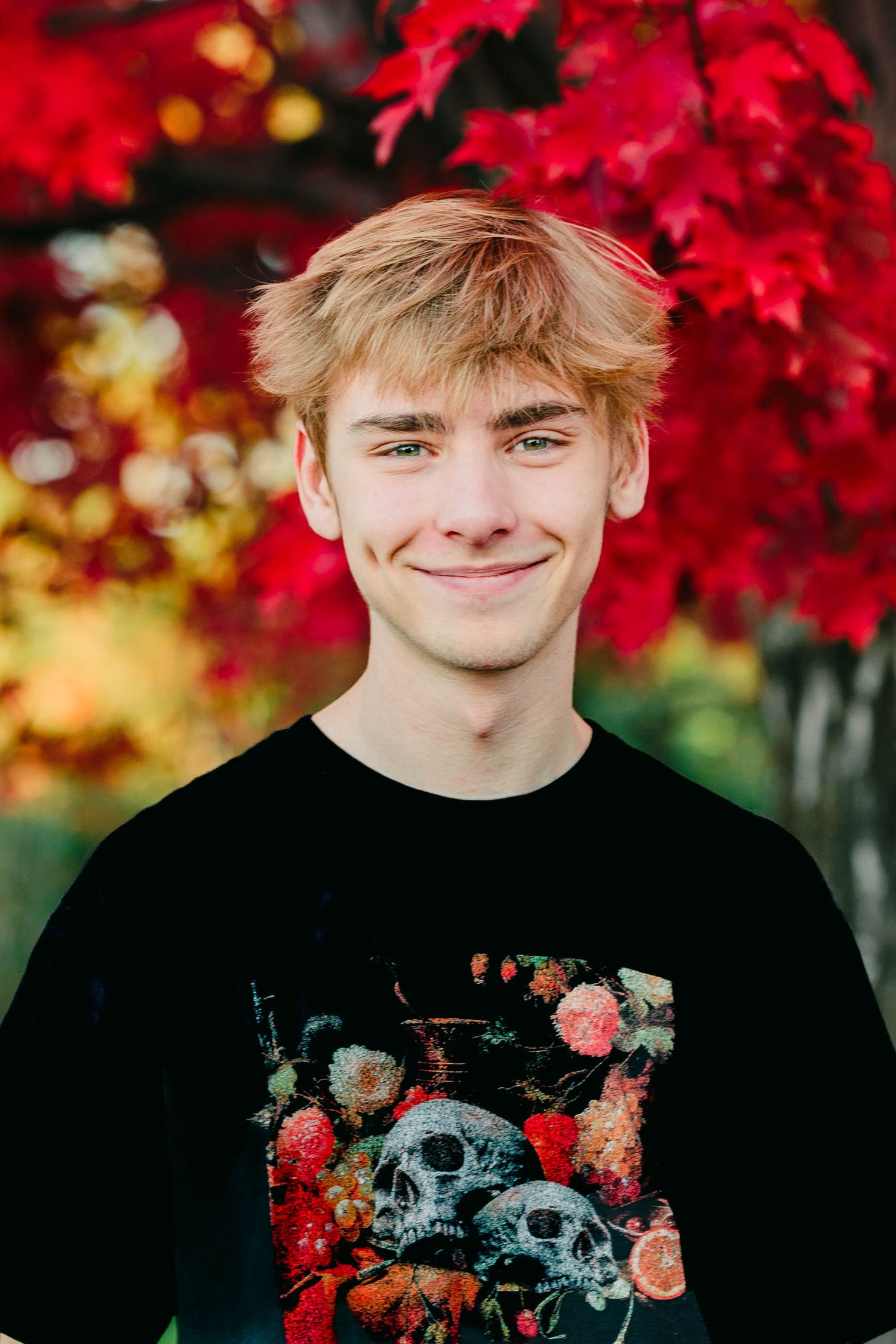Smiling teenage boy with blonde hair wearing skull graphic tee against red autumn leaves.