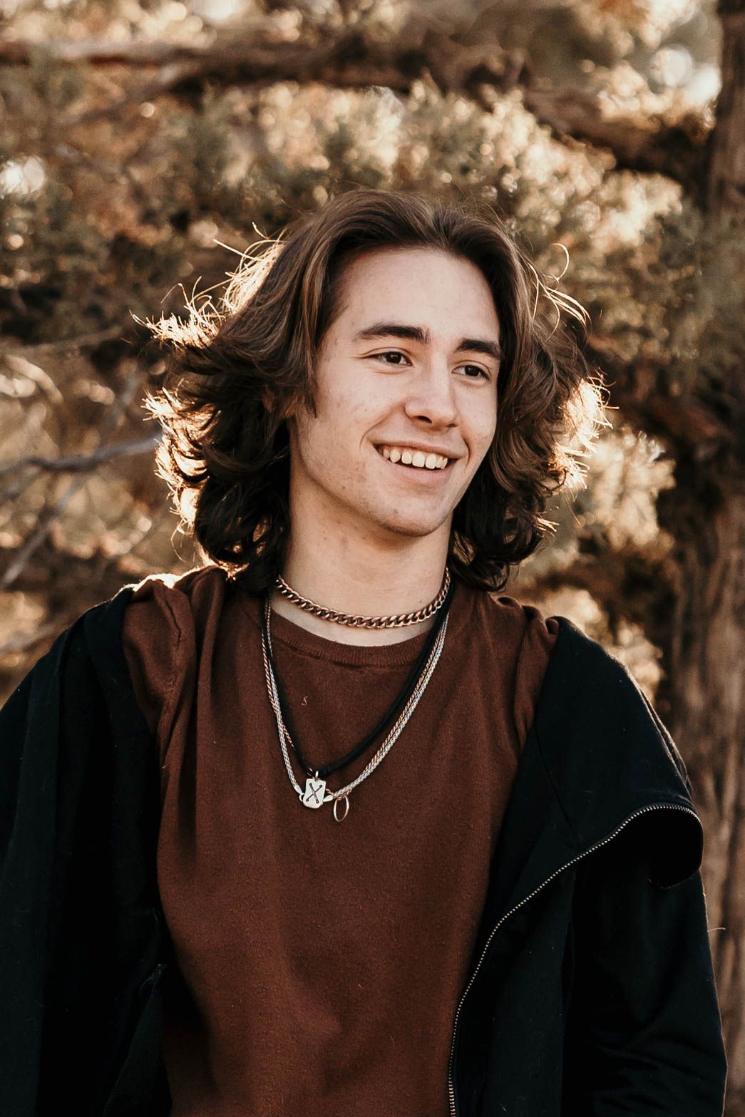 Smiling young man with long curly brown hair wearing a brown shirt and chain necklace outdoors.
